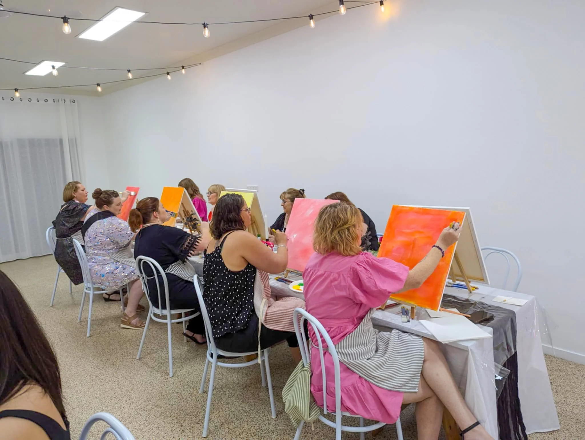 Group of women participating in a paint and sip class, painting canvases with warm orange and pink tones in a well-lit room with string lights.