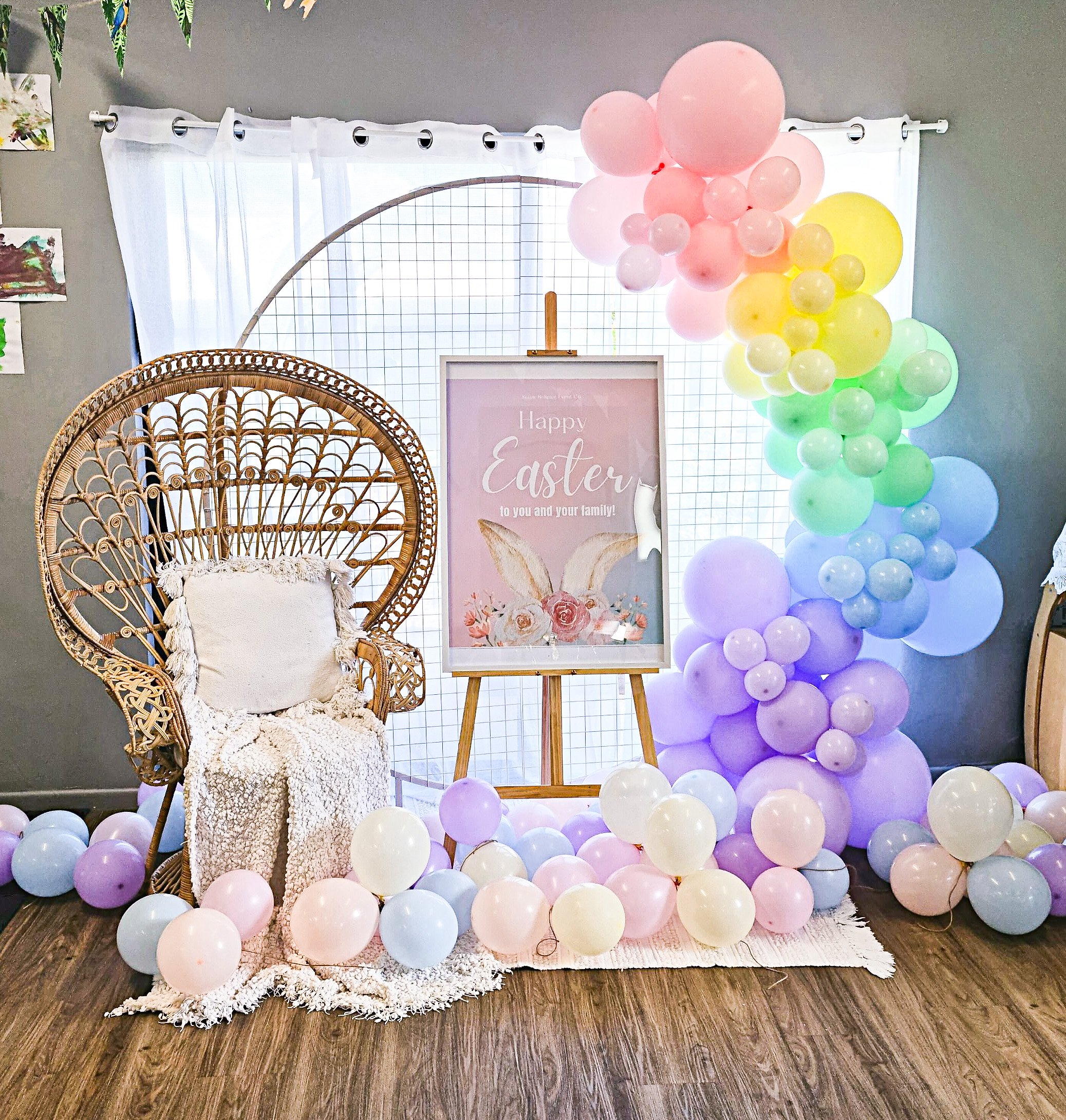 Decorative Easter display with a rattan peacock chair, a pastel-colored balloon arch, and a sign that reads, "Happy Easter to you and your family!" in front of a window with white curtains.