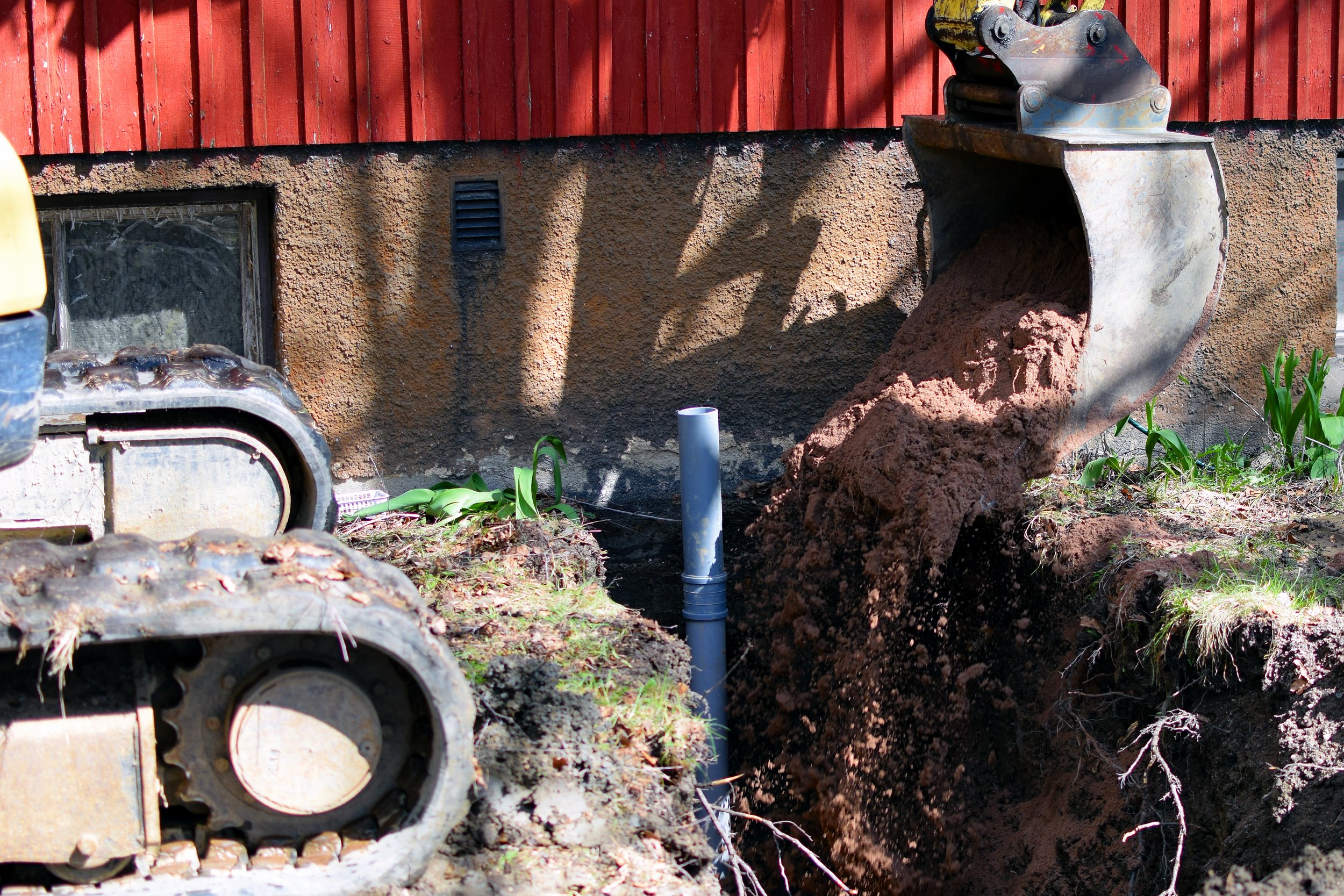 Excavator digging a trench outside a building with red siding.