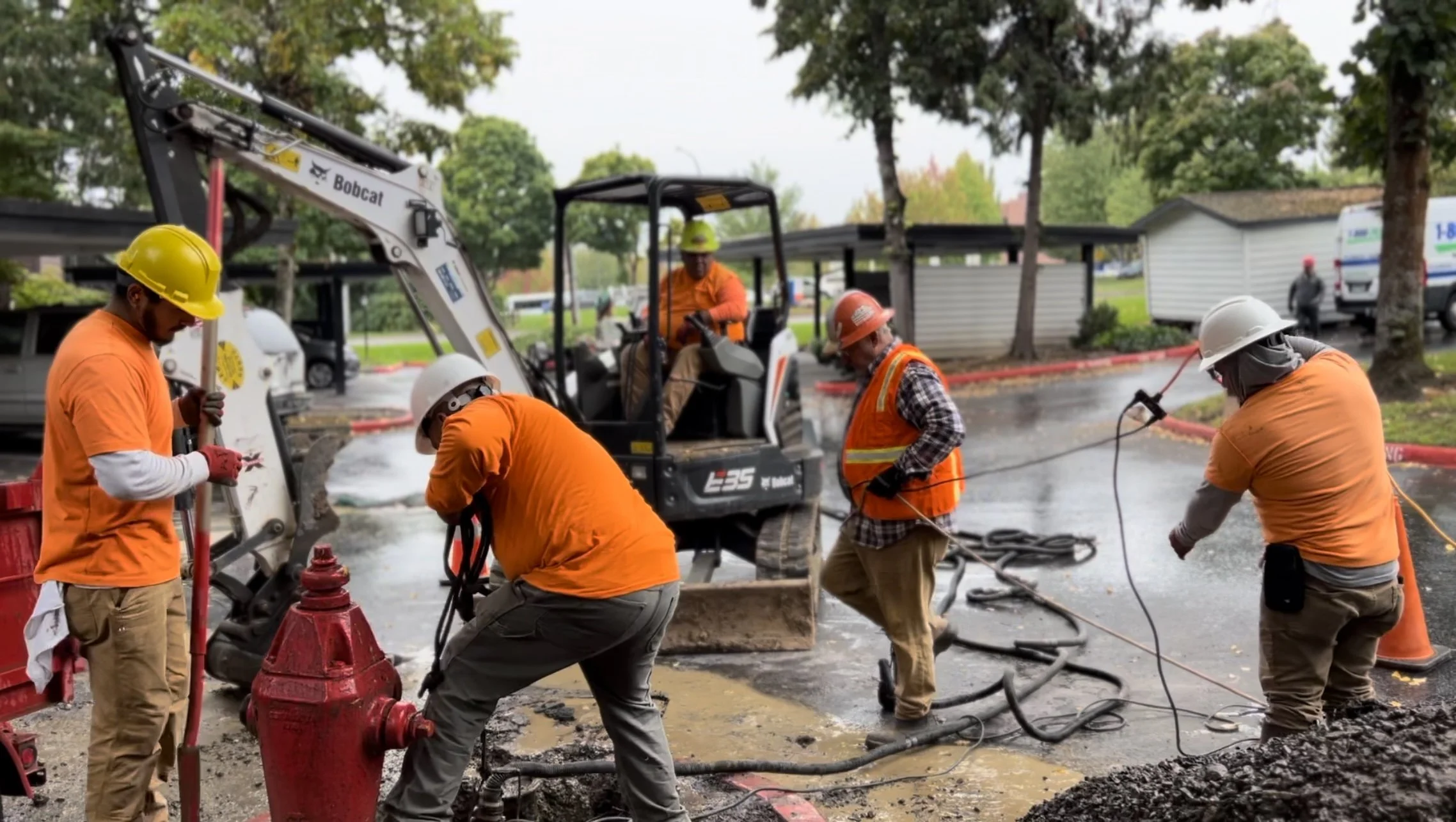 Construction workers in orange shirts and hard hats working on road repair, with a small excavator and various tools, in a wet outdoor setting.