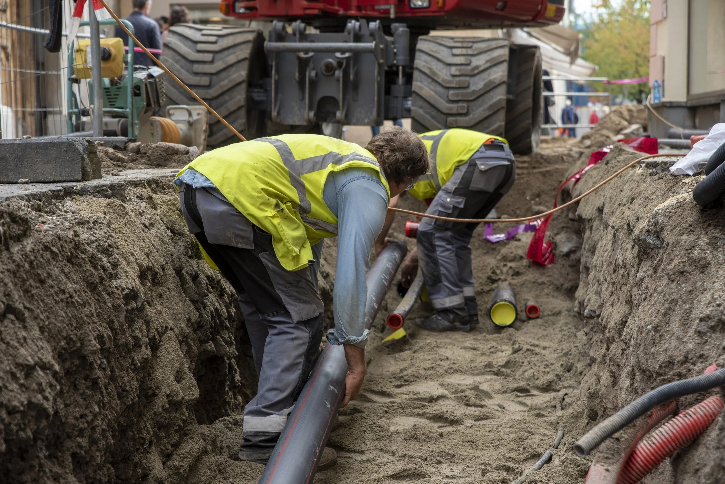 Workers in safety vests installing underground pipes in a construction trench, with a large vehicle in the background.