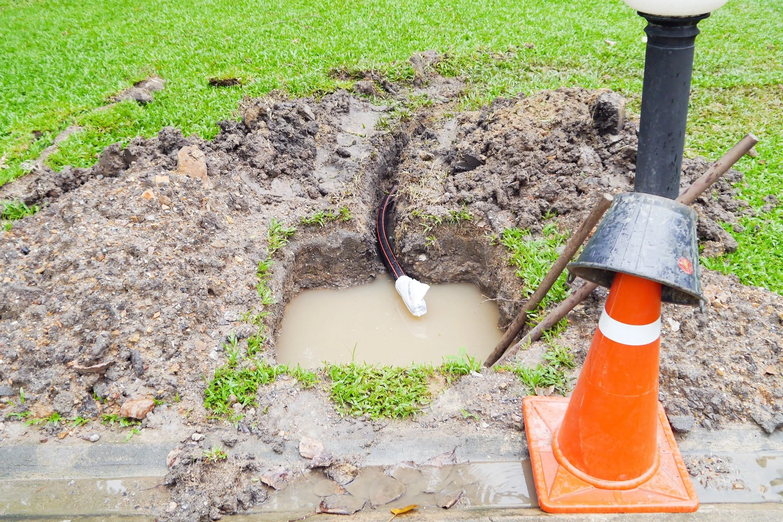 A hole in the ground with underground electrical cables exposed, surrounded by dirt and grass. An orange traffic cone and a black lamp post are nearby.