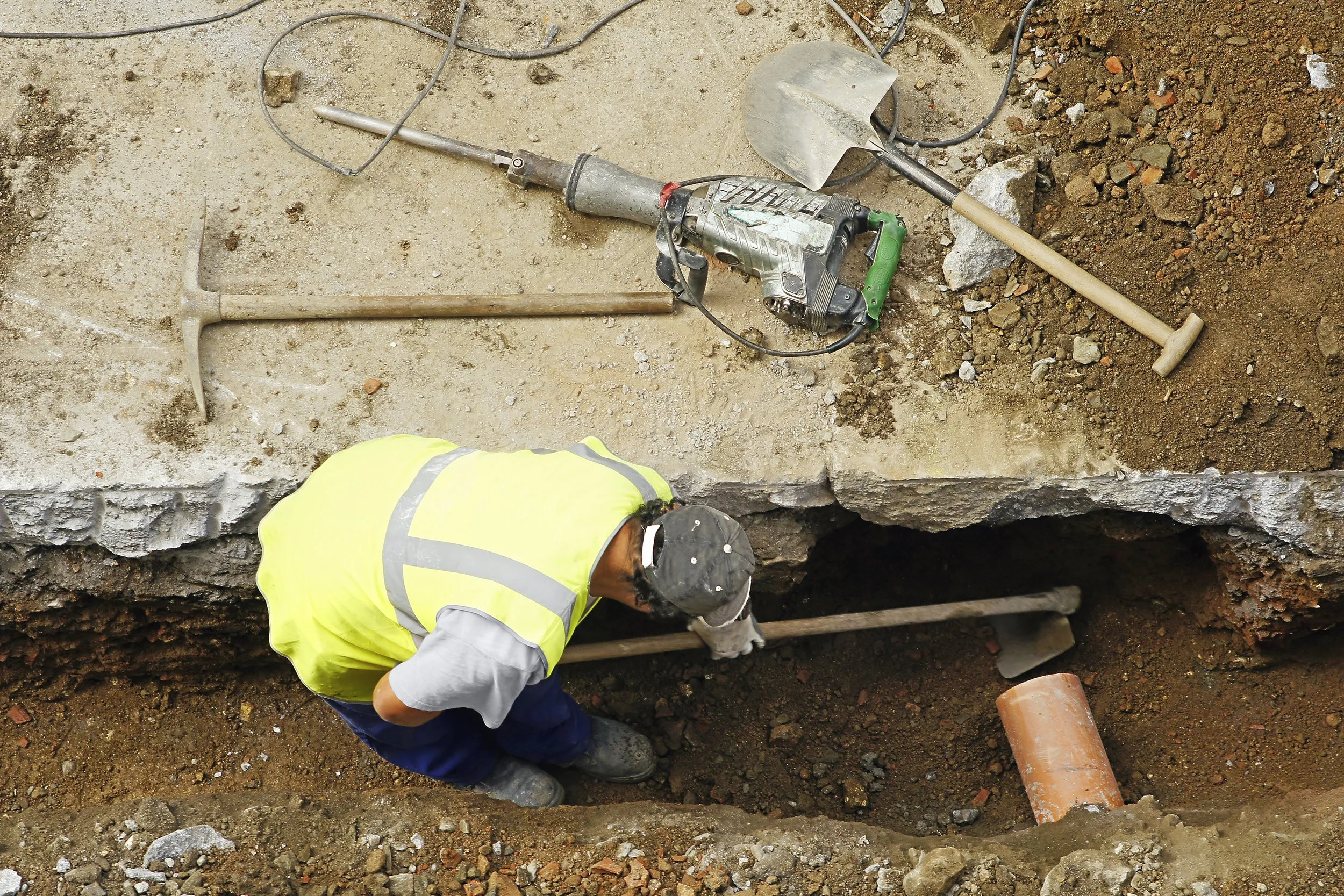 Construction worker in safety vest and helmet working in a trench with tools and machinery nearby.