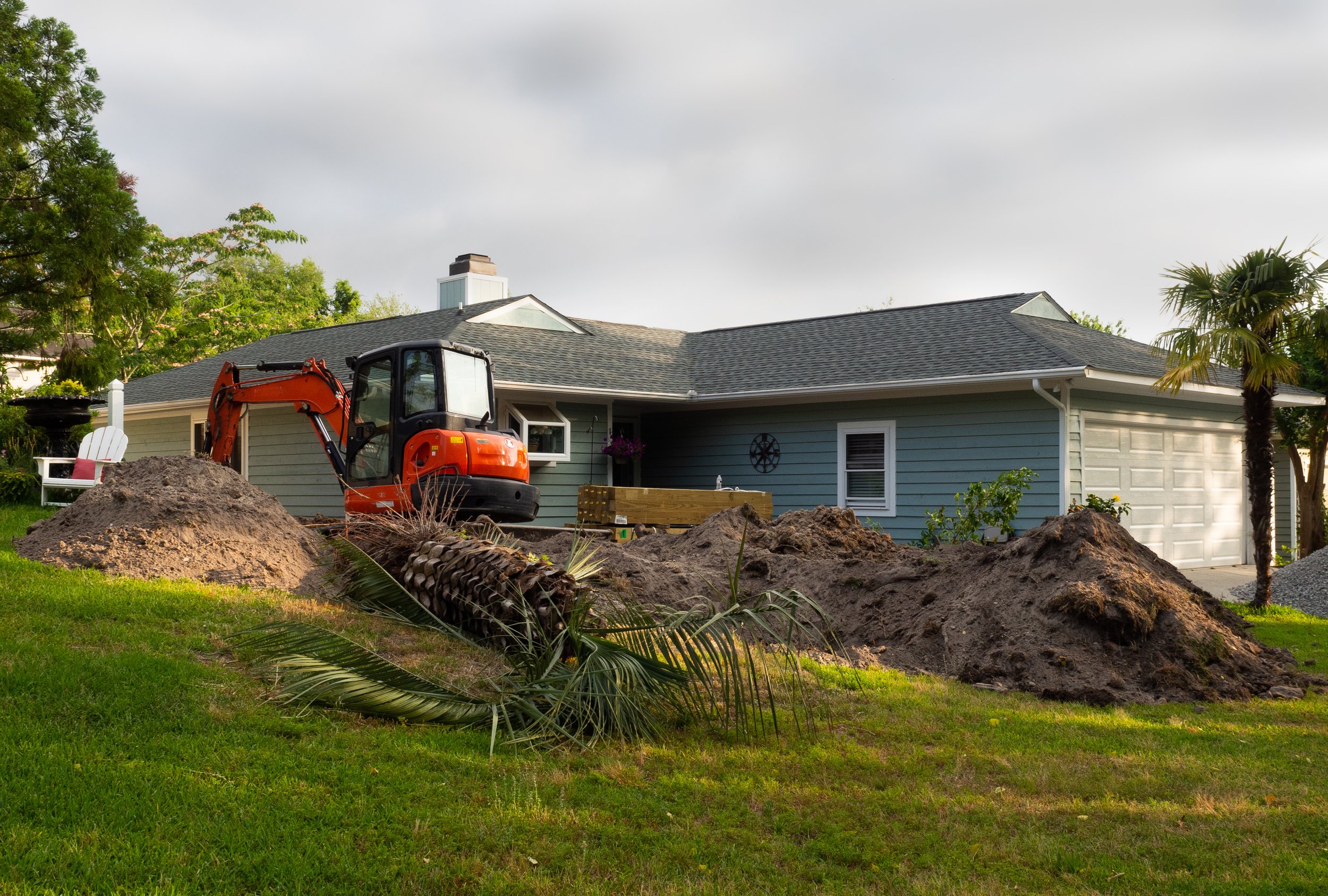 Construction work in progress at a residential backyard, with an orange mini excavator and dug-up soil around a house with blue siding, white trim, and a garage door, surrounded by green grass, trees, and landscaping.