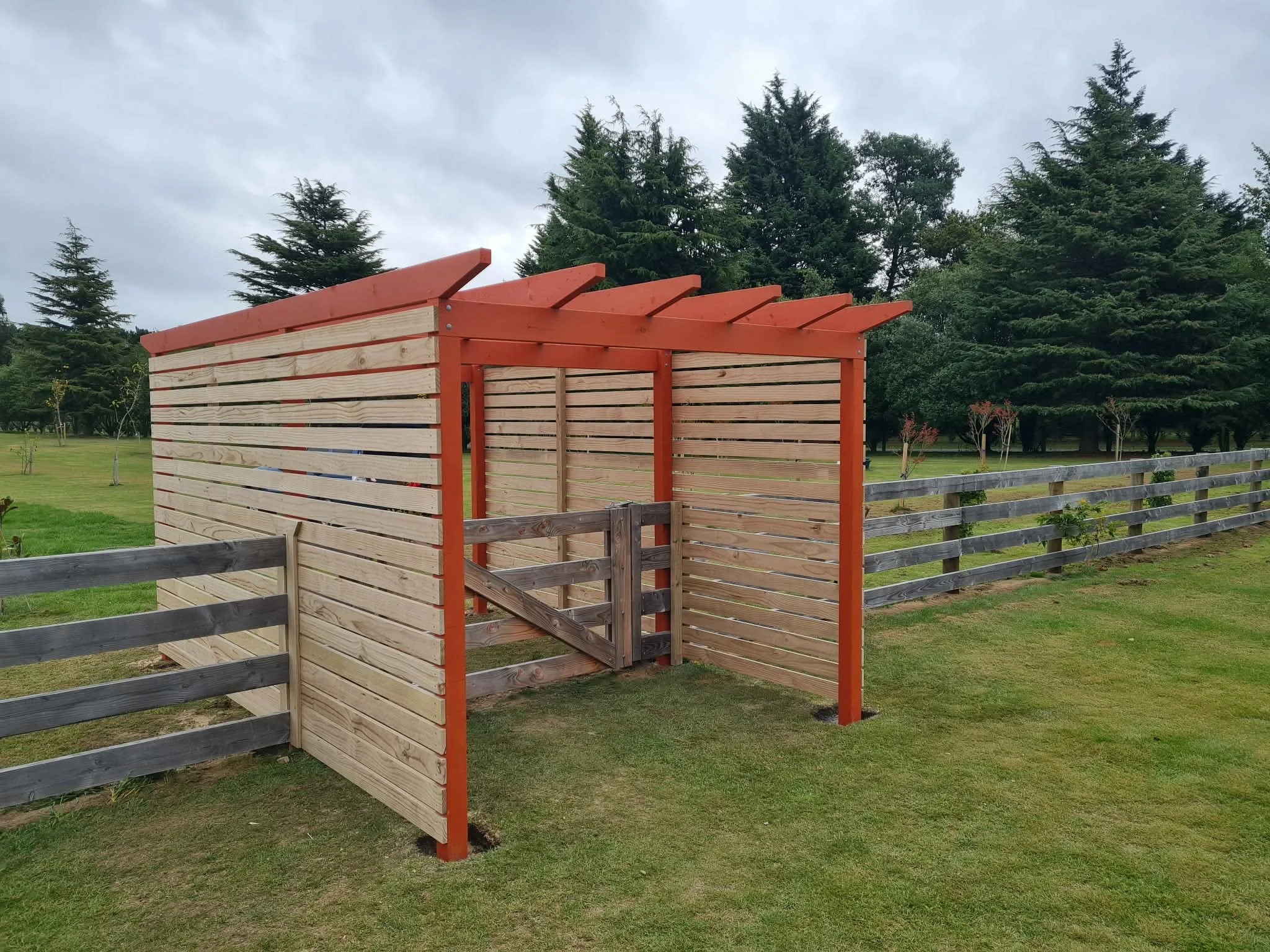 A wooden spiritual entraceway with horizontal slats, painted red trim, and a small gate, situated on a grassy area with trees in the background.