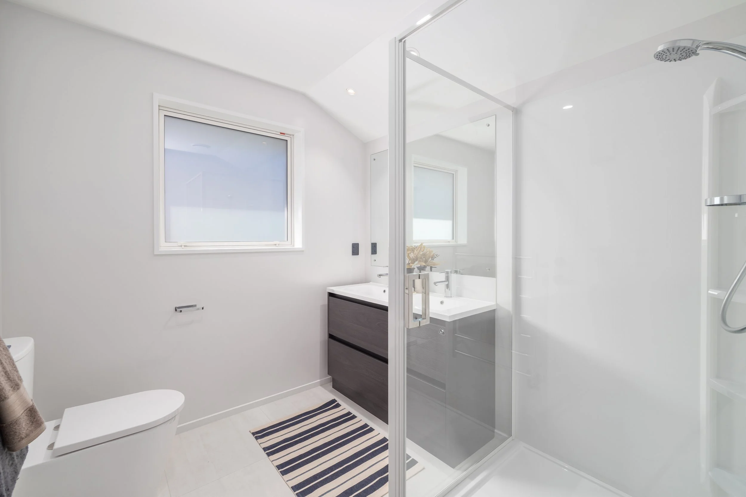 New Home Building, Modern bathroom with white walls, a window, a glass-enclosed shower, a dark vanity with a white sink, a mirror, a striped rug, and a toilet.
