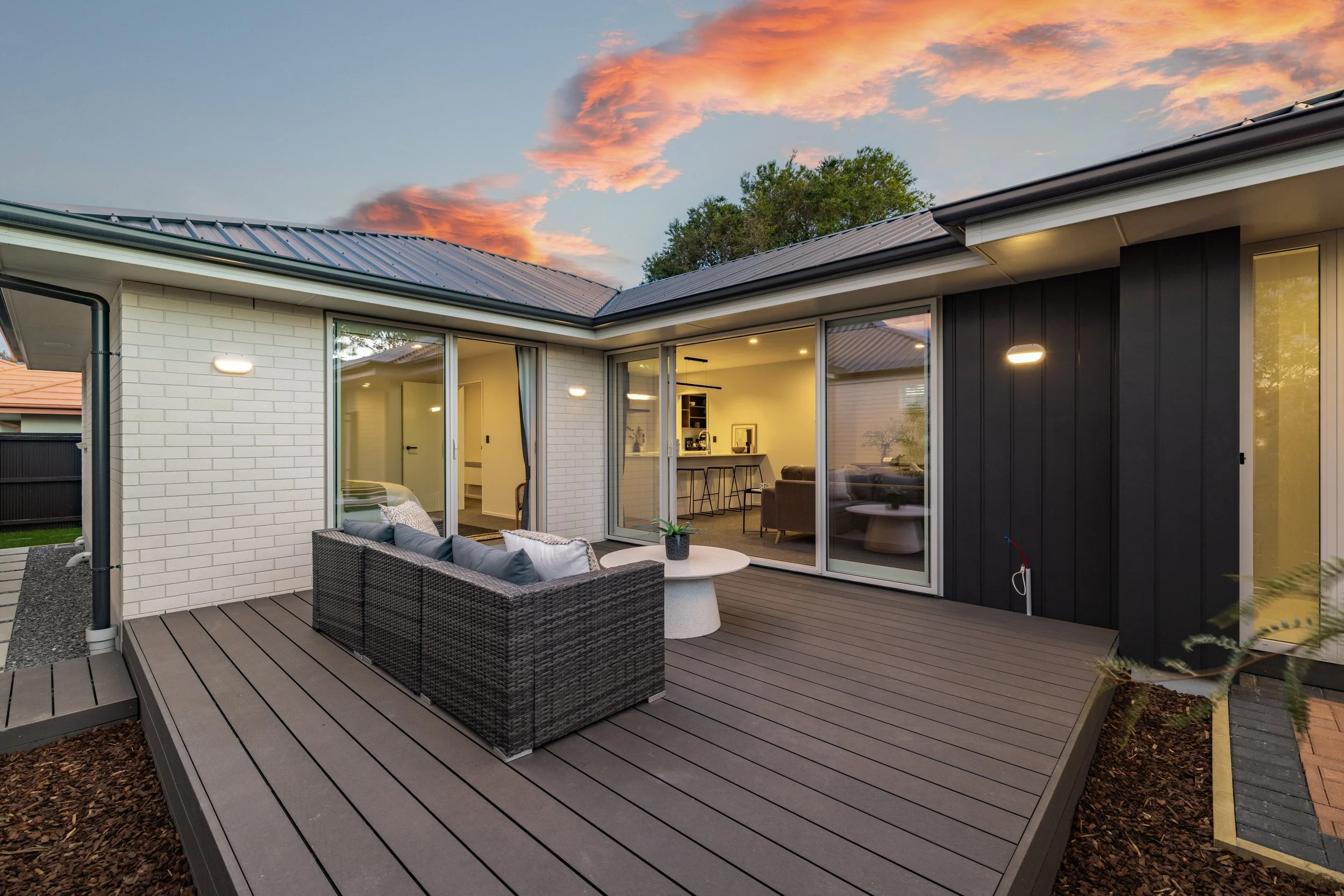 Modern house exterior with a spacious wooden deck, wicker outdoor sofa, sliding glass doors showing lit interior, during sunset.