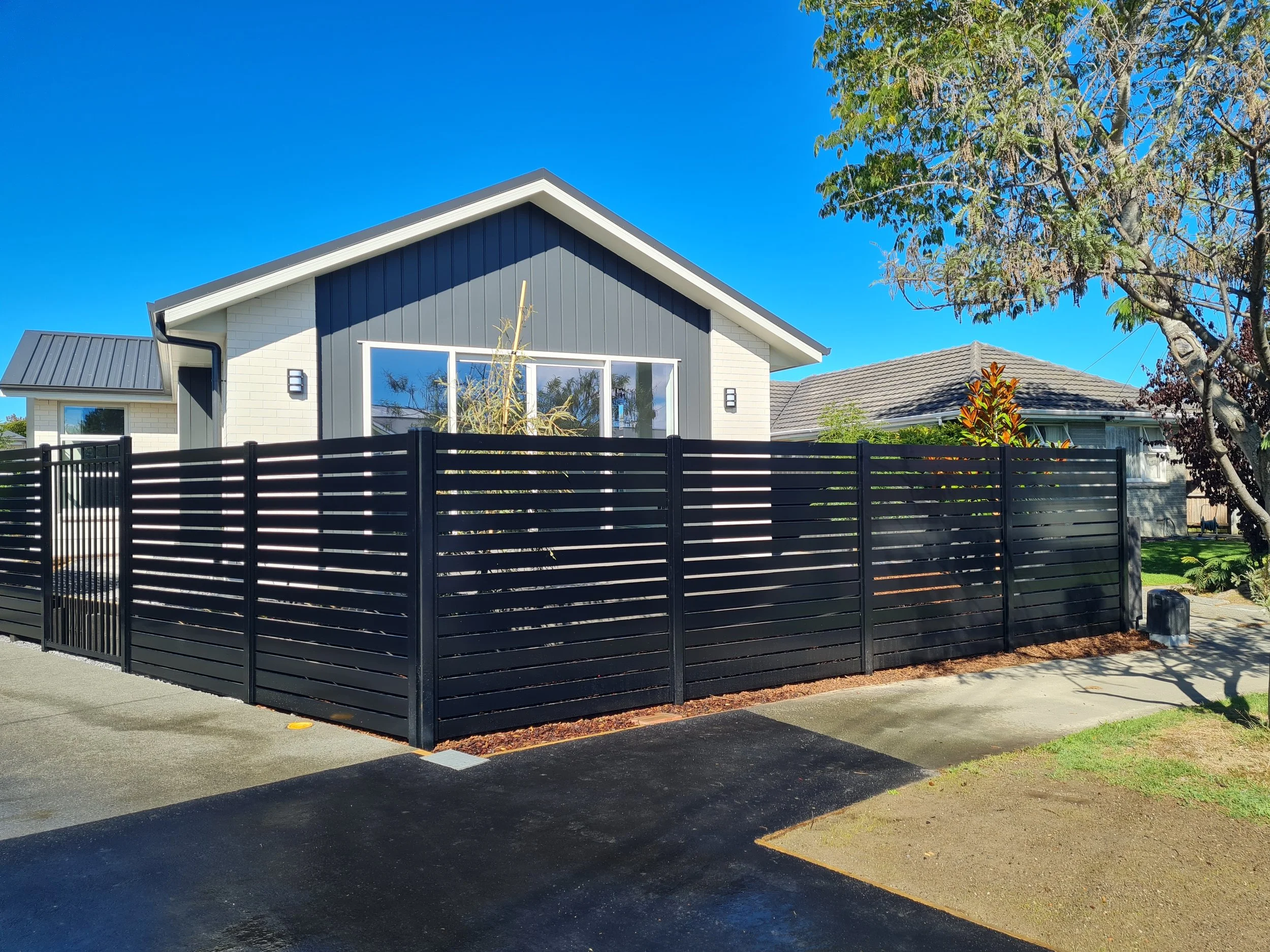 Modern house with white brick walls, dark blue vertical siding, large windows, and a black horizontal slat fence in the front yard. Clear blue sky and trees in the background.