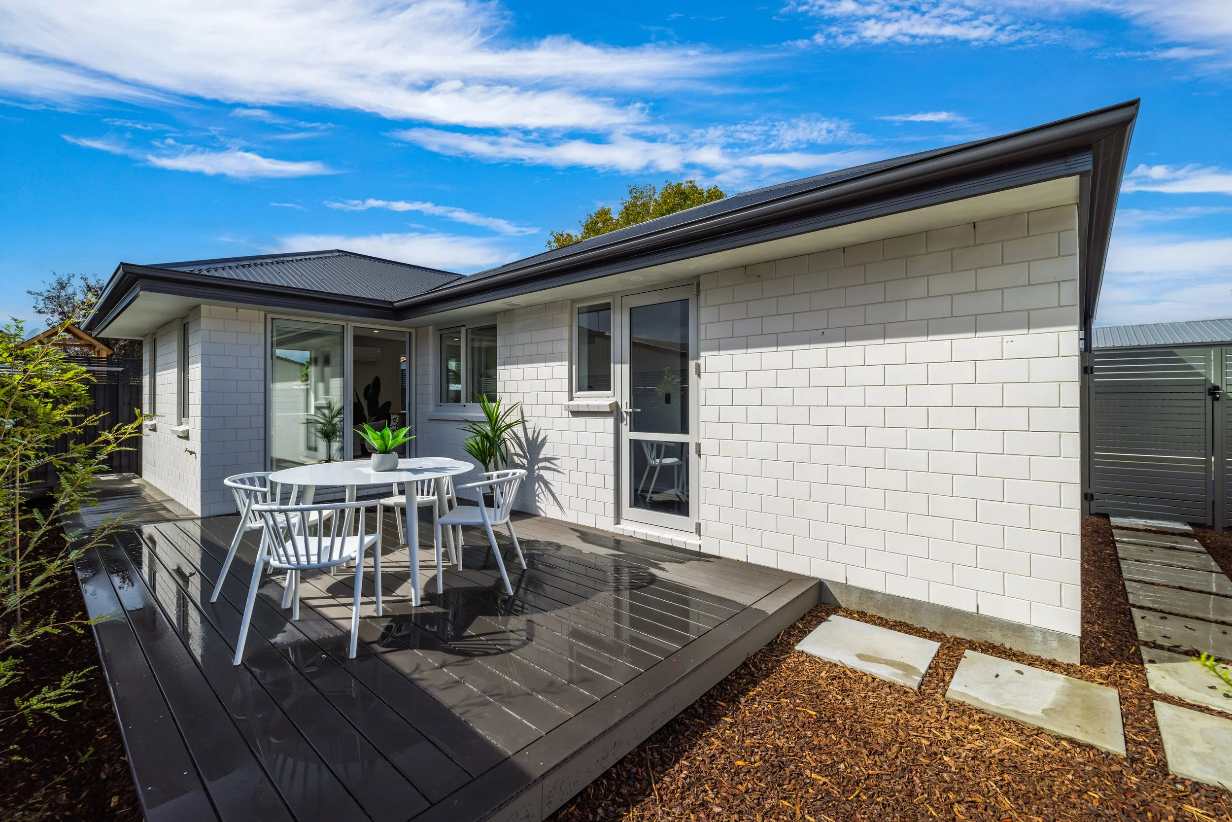 Modern white brick house with a small outdoor deck made of sustainable, no maintenance material, table with four white chairs, potted plants, and a sliding glass door under a partly cloudy blue sky.
