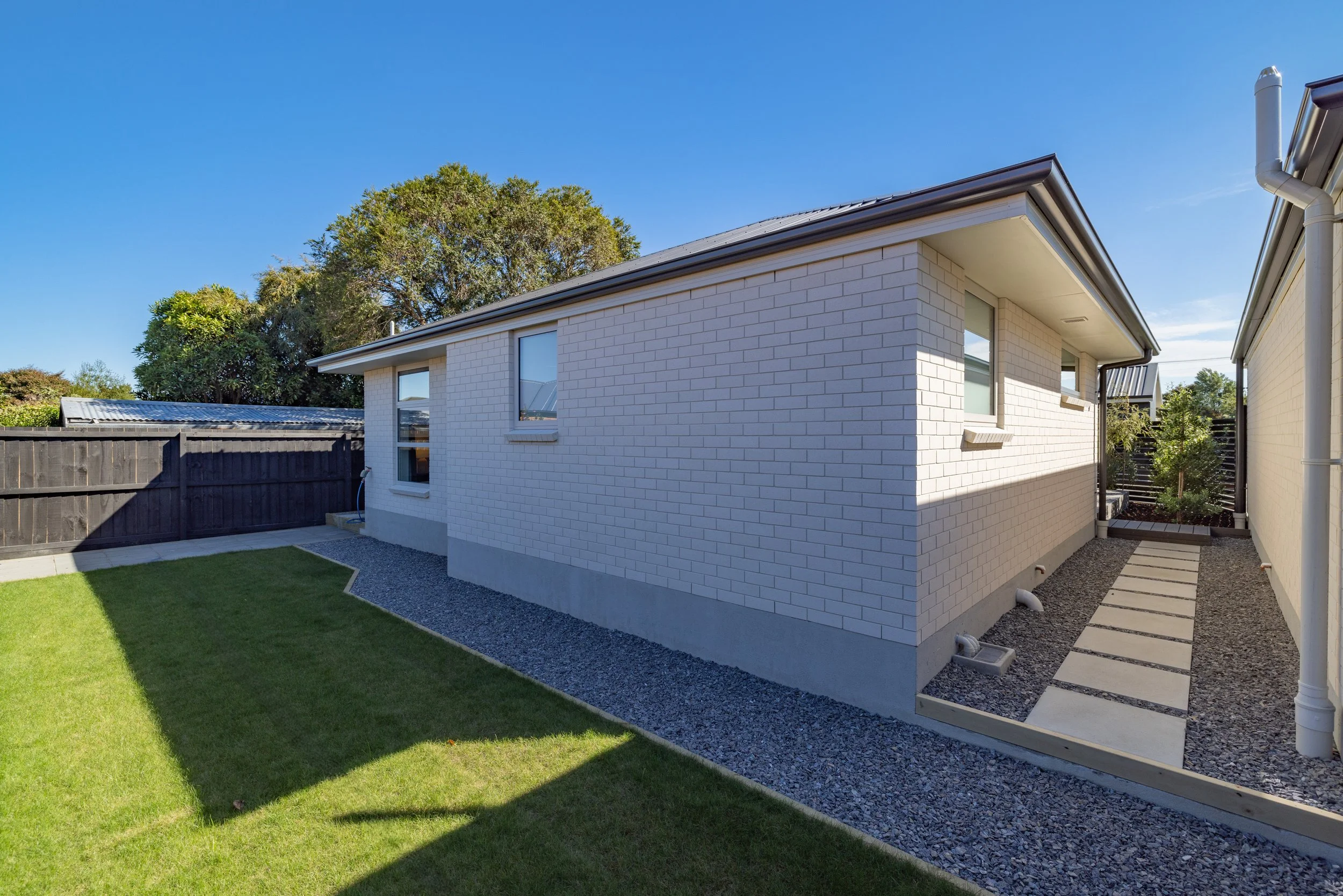 Backyard view of a modern white brick house with a small lawn, gravel pathway, and a wooden fence under a clear blue sky.