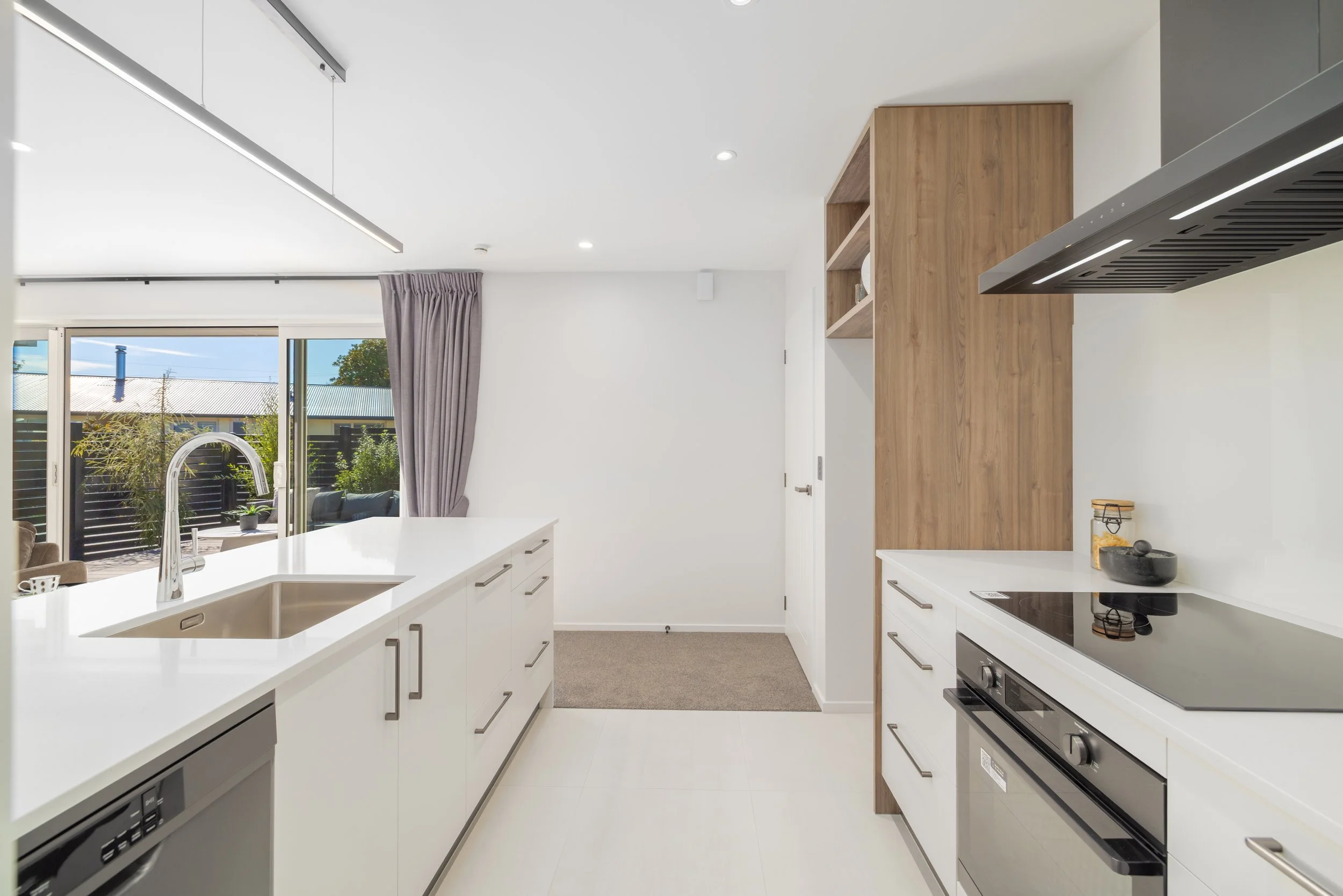 New Home Christchurch, Modern kitchen with white island countertop, stainless steel sink, black stovetop, wooden cabinetry, large glass sliding door, gray curtains, and outdoor patio view