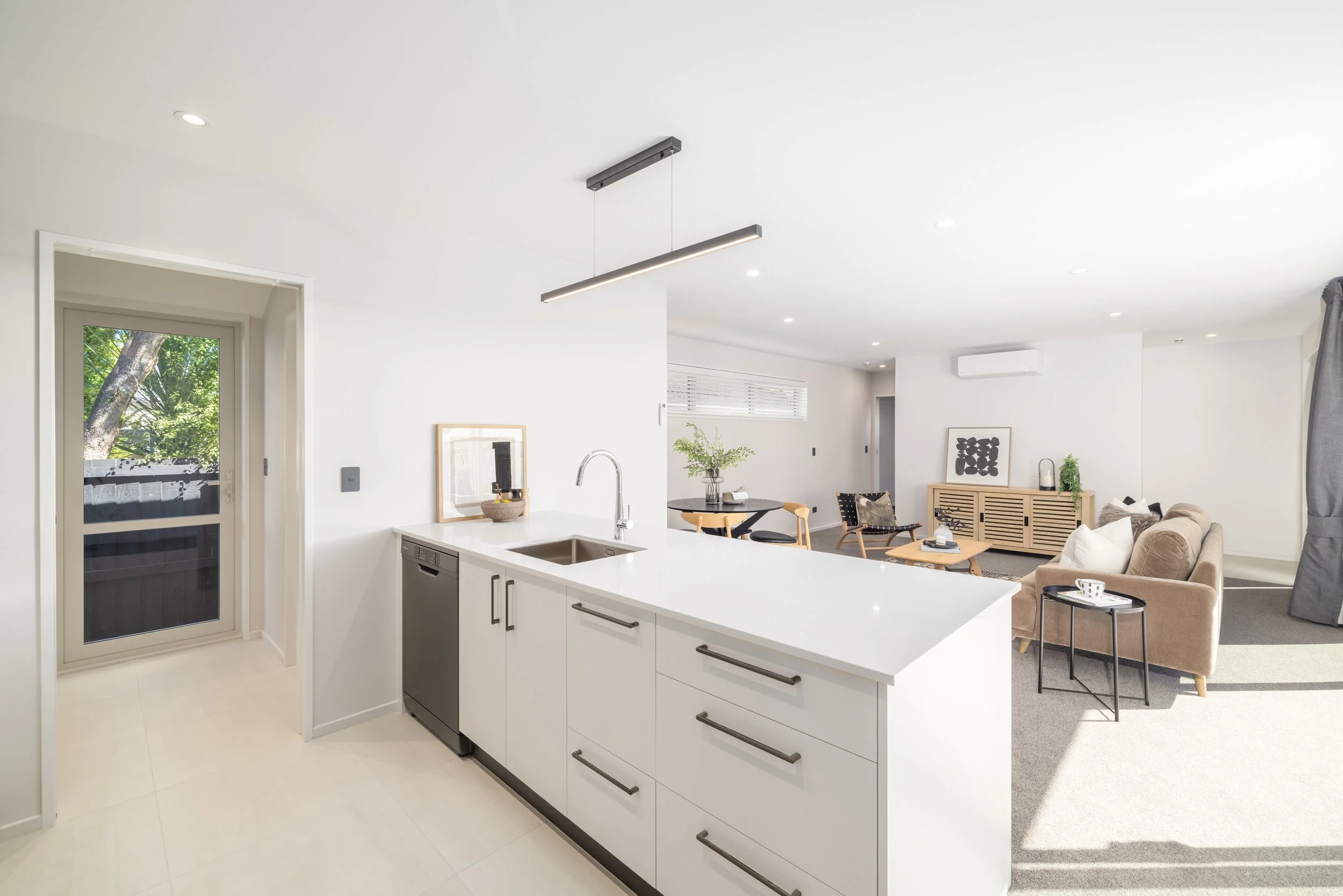 Modern open-plan living room and kitchen with white walls, beige sofa, black and wooden furniture, and large windows.