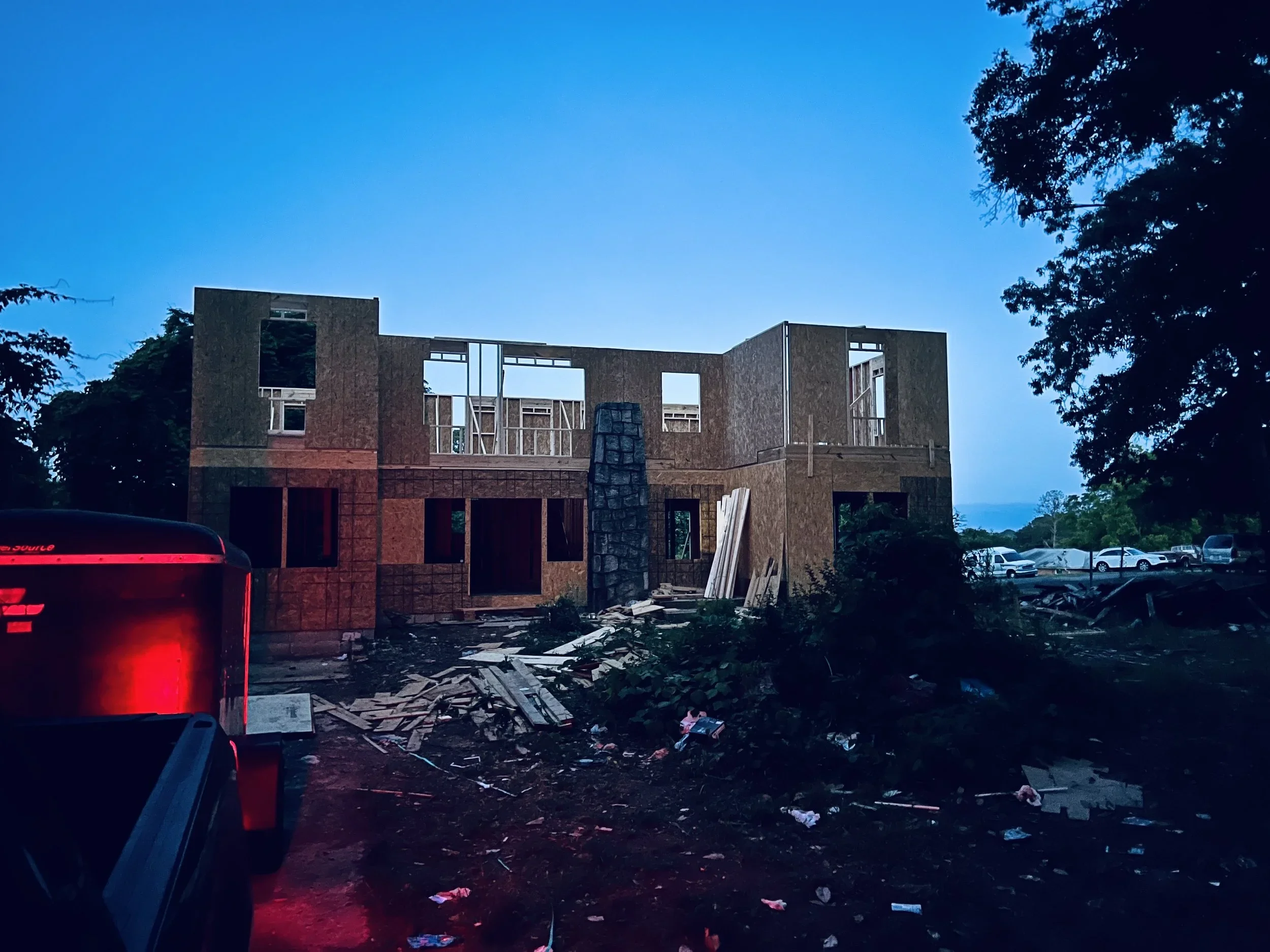 A house under construction with wooden framing and partially installed walls, surrounded by construction debris and parked cars at dusk.