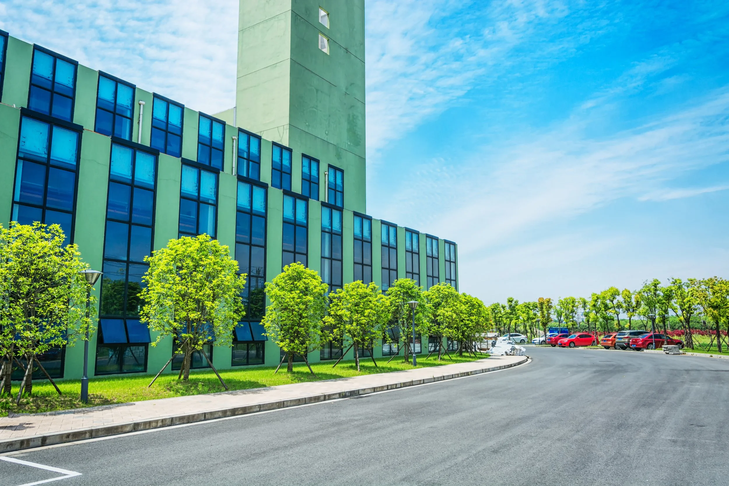 A modern green building with blue-tinted windows, a curved asphalt driveway, green trees, and parked cars under a partly cloudy sky.