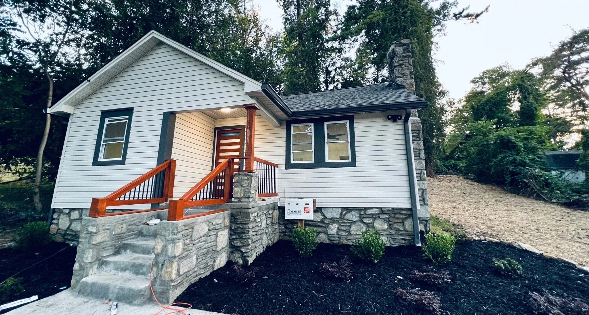 Newly constructed small house with white siding, stone foundation, and a front porch with wooden steps and railing, surrounded by landscaping and trees.
