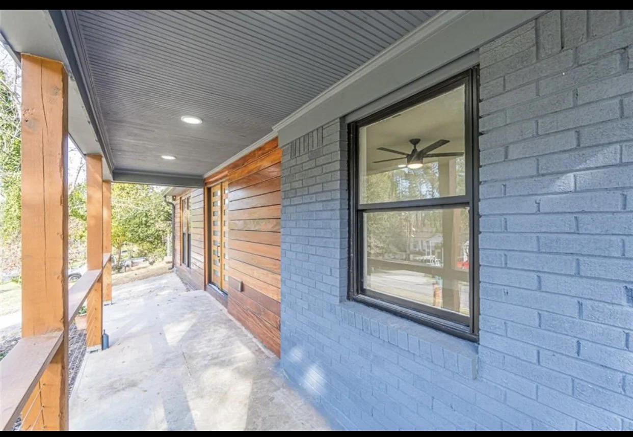 Porch area of a house with gray painted brick wall, a window with black frame, wooden wall siding, and unfinished concrete floor.