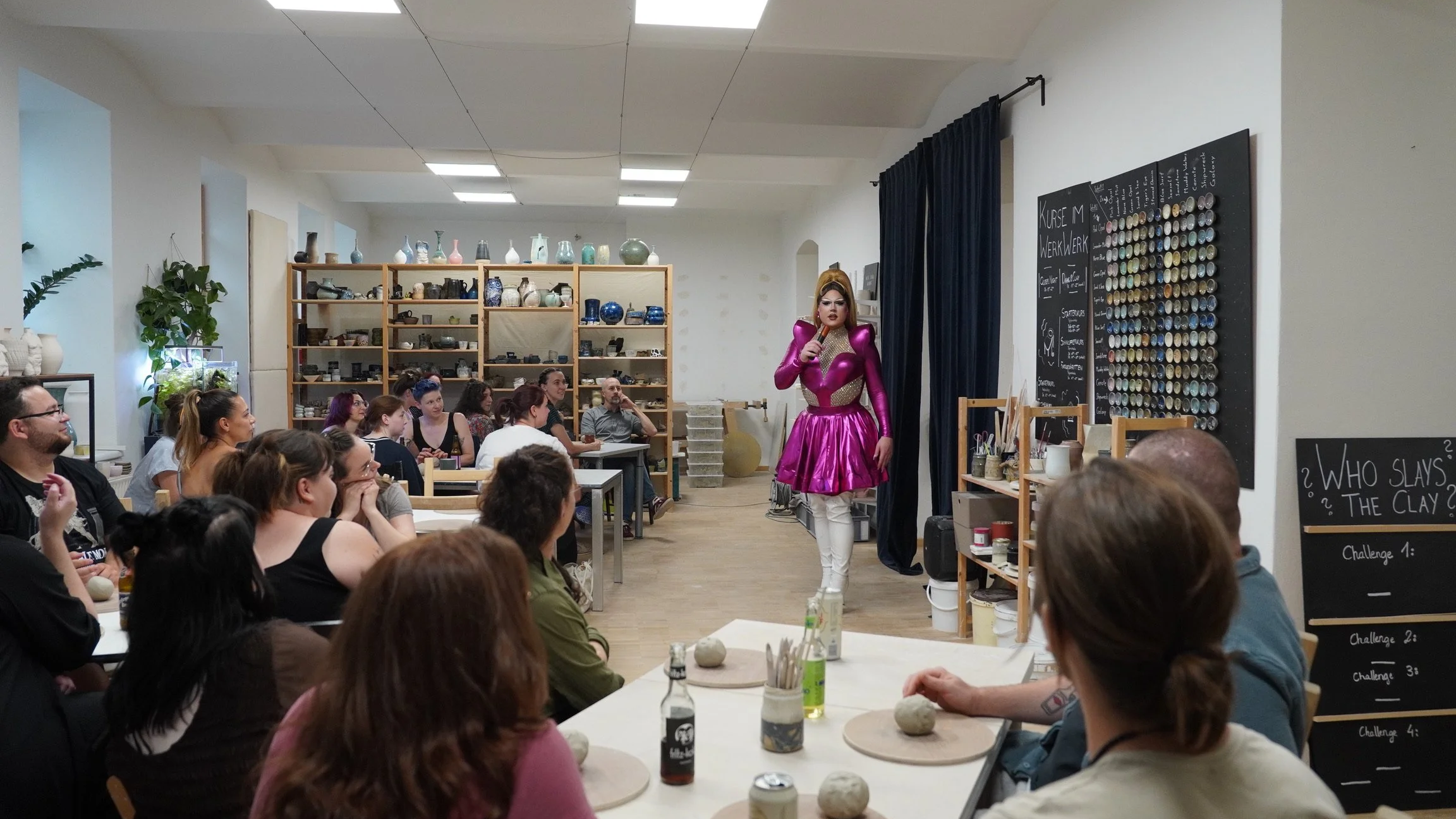 Eine Frau in einem pinkfarbenen Kleid und weißen Stiefeln steht mit einem Mikrofon vor einer Gruppe von Menschen in einem Kreativraum. Im Hintergrund sind Regale mit Keramik und Kunstmaterialien, und auf der rechten Seite sind Tafel mit Kursinformati
