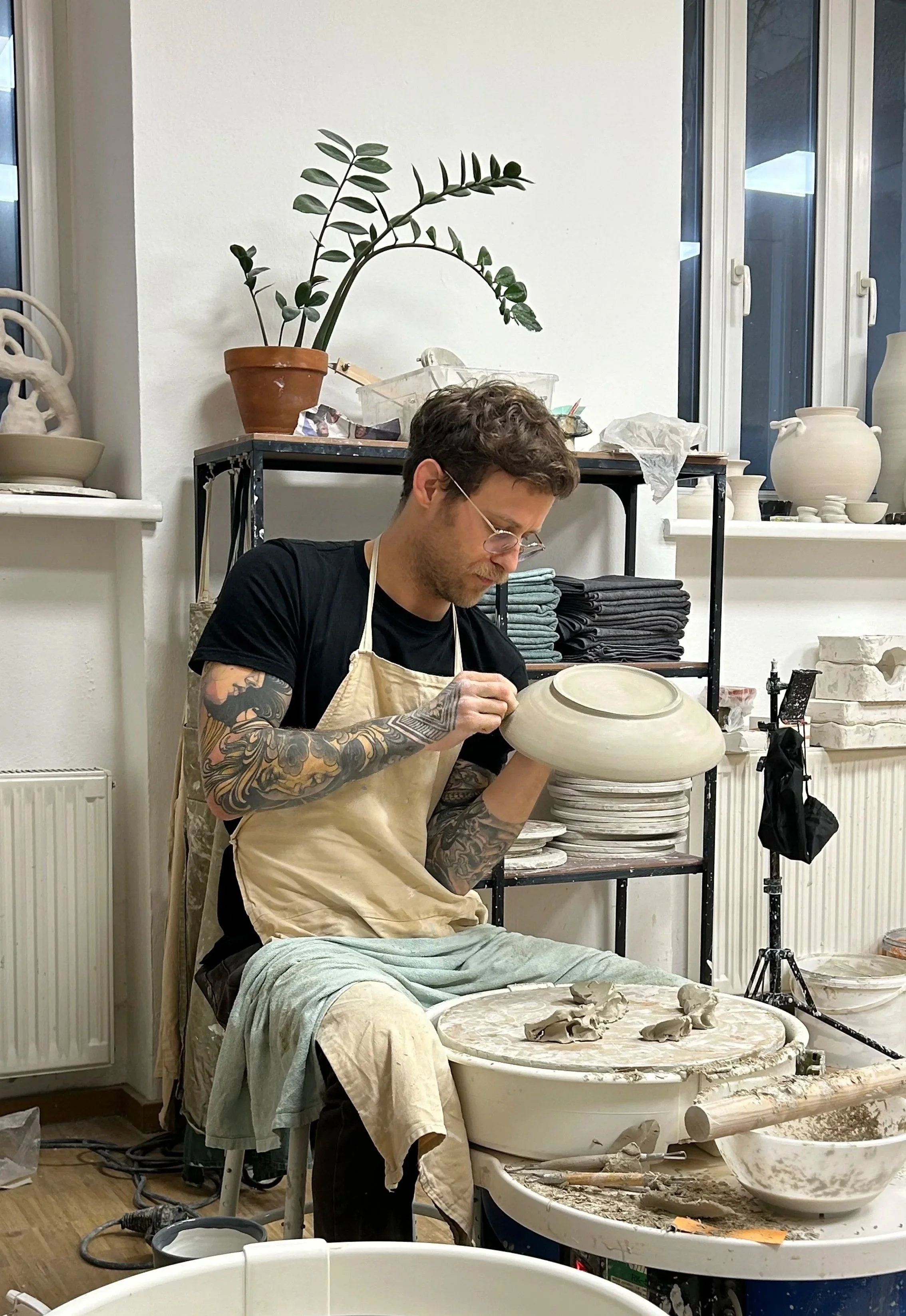 Ein Mann mit Tattoos arbeitet in einem Keramikstudio an einer Töpferscheibe, auf der er eine große, flache Tonschale formt.