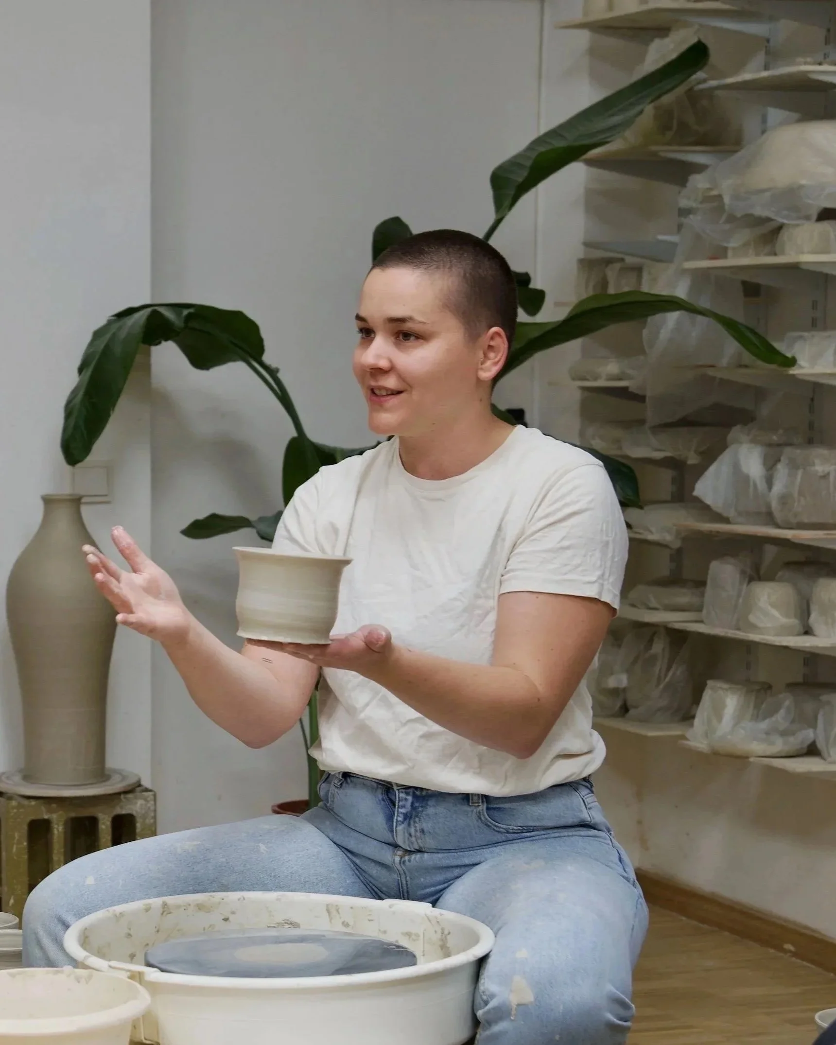 Junge Frau in Keramikatelier, hält eine geformte Keramikschale in den Händen, während sie an ihrem Werk arbeitet, mit Töpferware und Werkzeugen im Hintergrund.