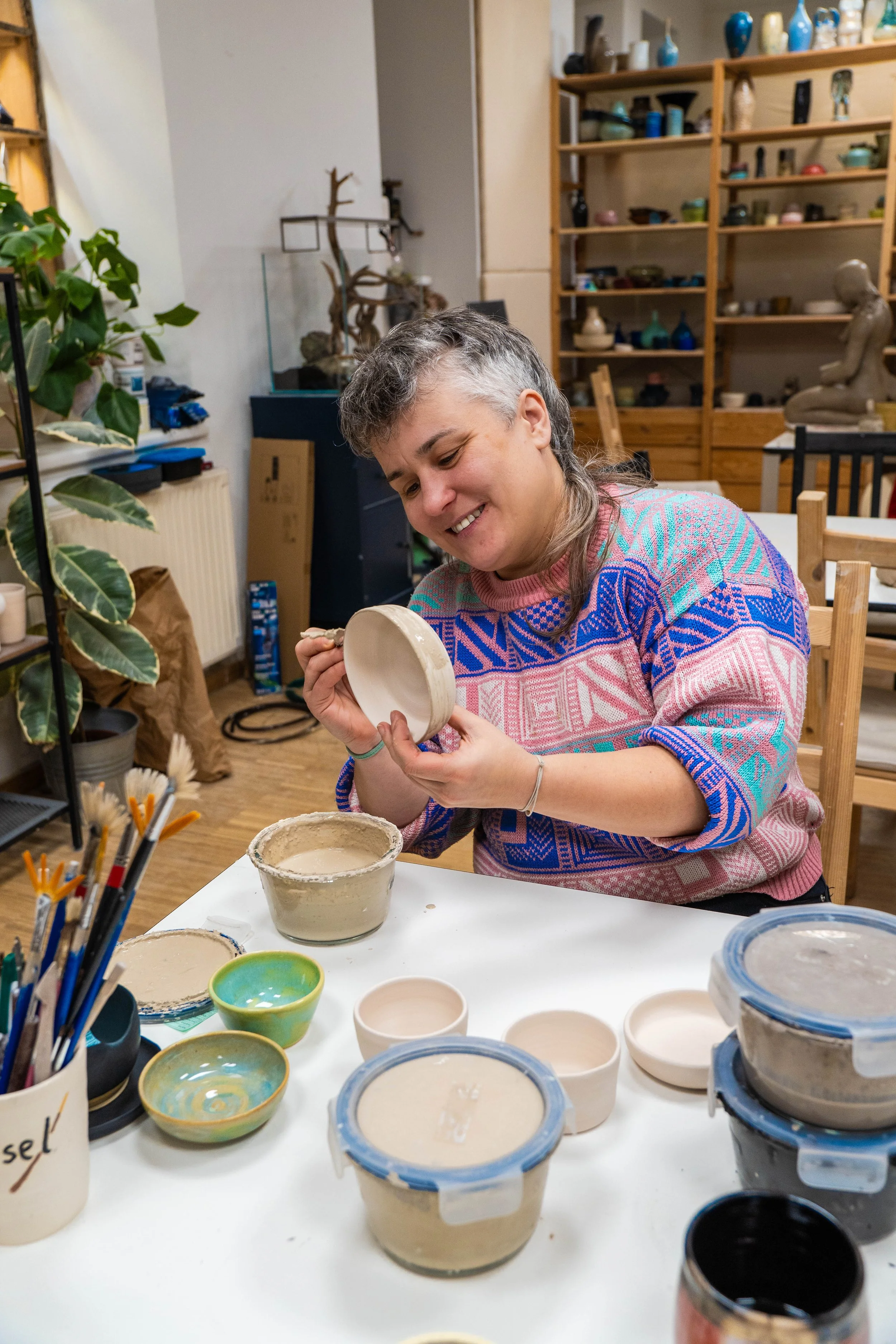 Eine Frau arbeitet mit Ton und bemalt eine Keramikschale in einem Workshop- oder Atelierraum, umgeben von Töpferwaren, Werkzeugen und Pflanzen.