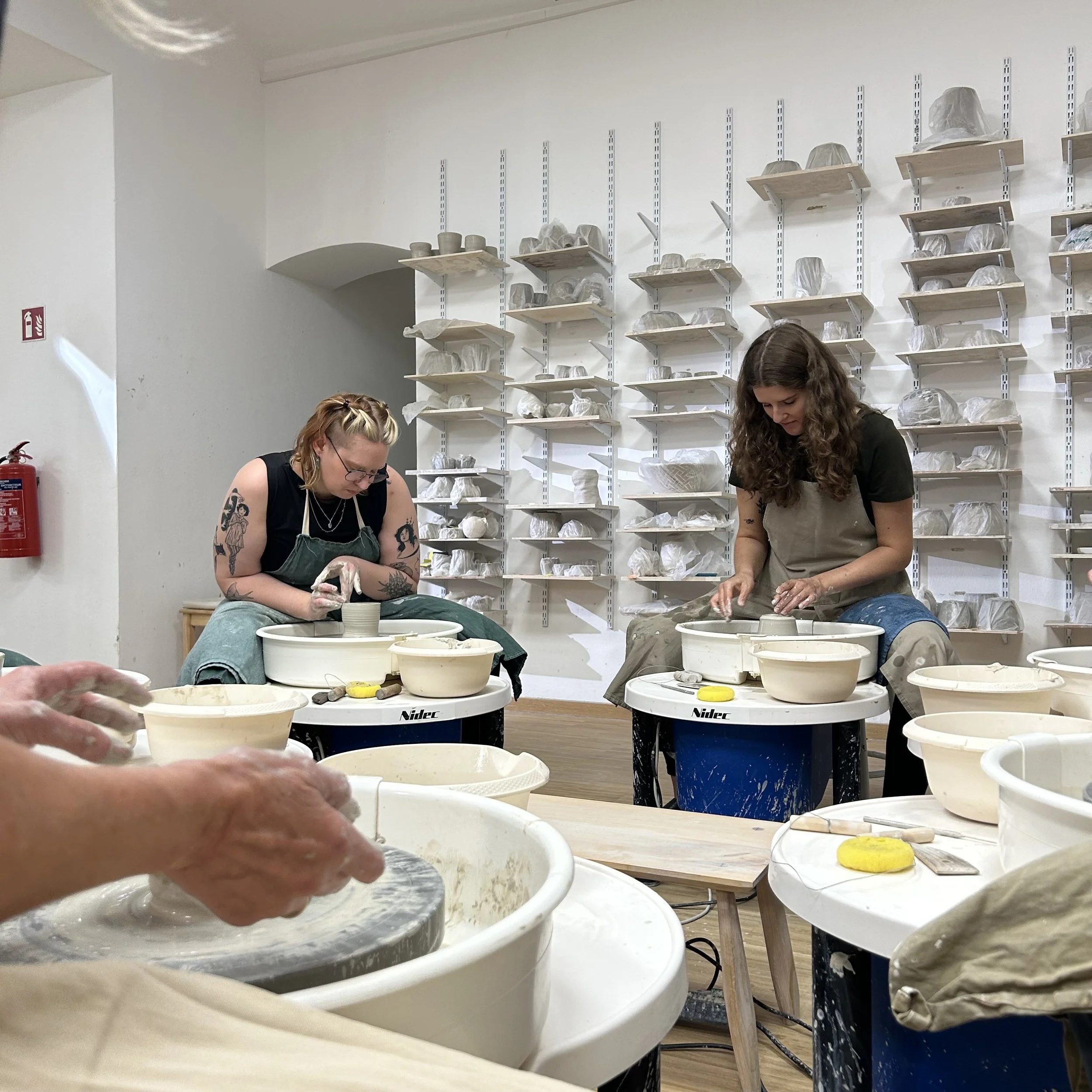 Zwei Frauen beim Töpfern in einem Keramikstudio, sitzend an Tischen mit Töpferdrehscheiben, mit mehreren Töpferwaren und Werkzeugen, an einer weißen Wand mit Regalen voller unvollendeter Keramikstücke.