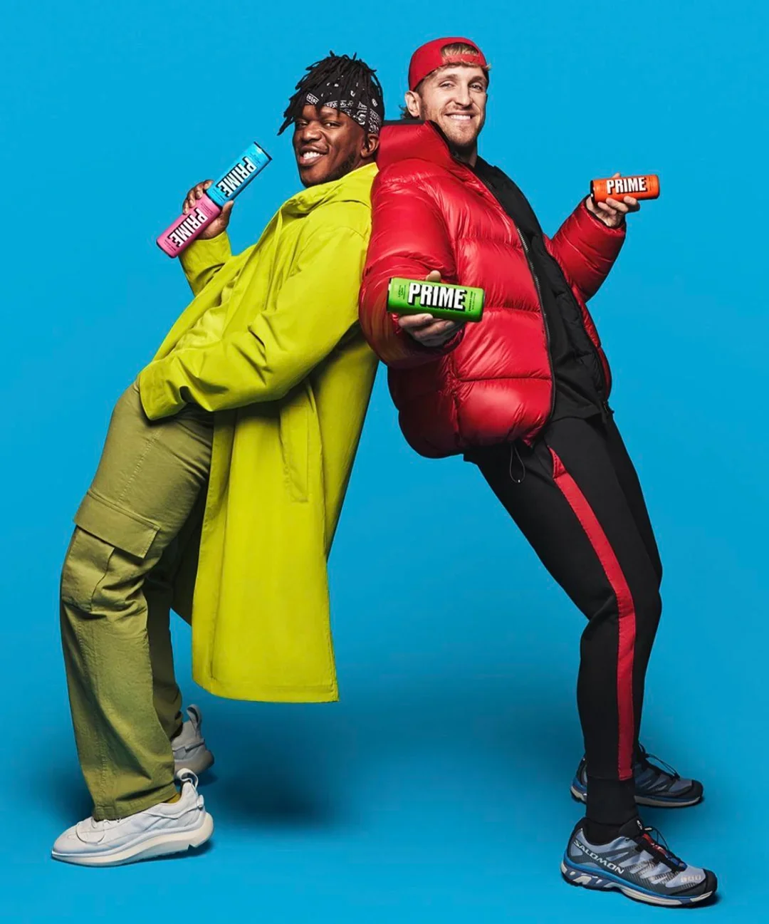 Two men smiling and standing back to back against a bright blue background, each holding a colorful Prime energy drink. One man wears a bright yellow jacket and beige pants, the other sports a red puffy jacket and black pants.