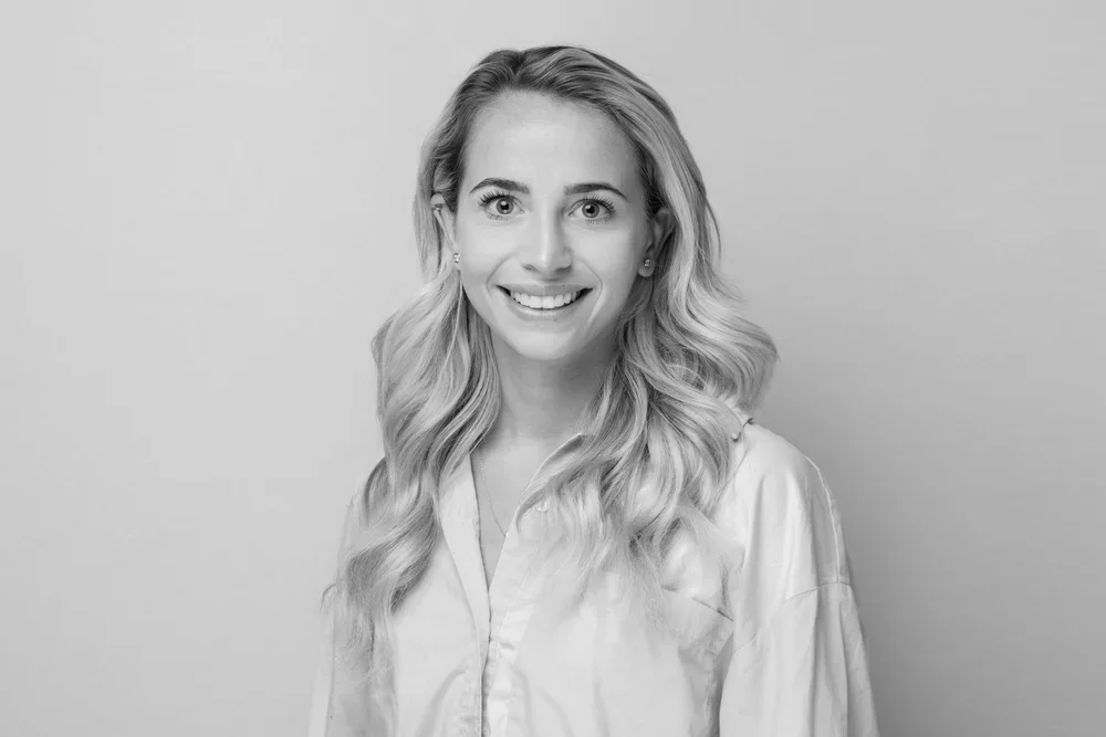 Black and white portrait of a smiling woman with wavy blonde hair, wearing a light-colored button-up shirt, standing against a plain light background.