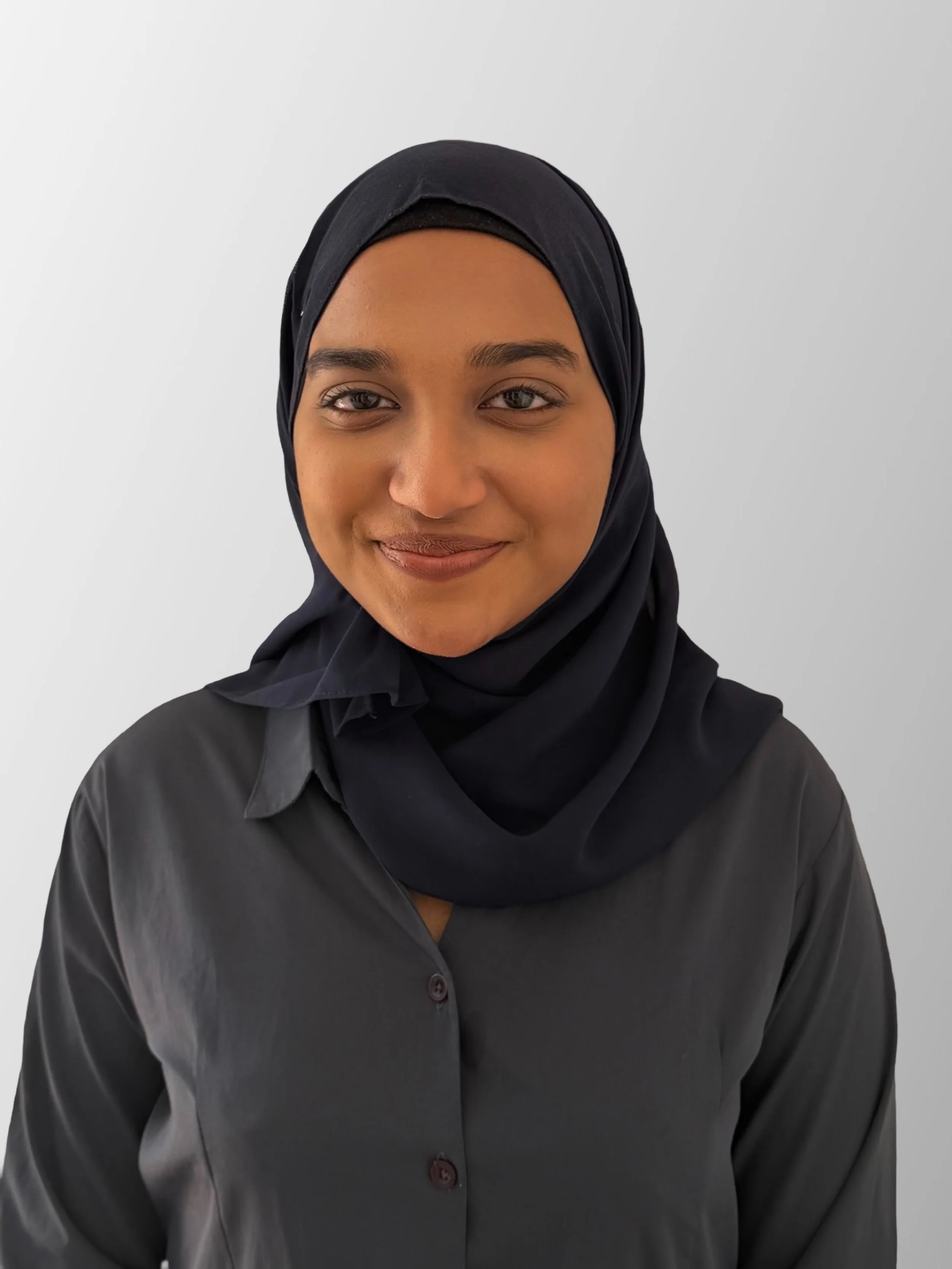 A young woman wearing a black hijab and black button-up shirt, smiling slightly, against a plain white background.