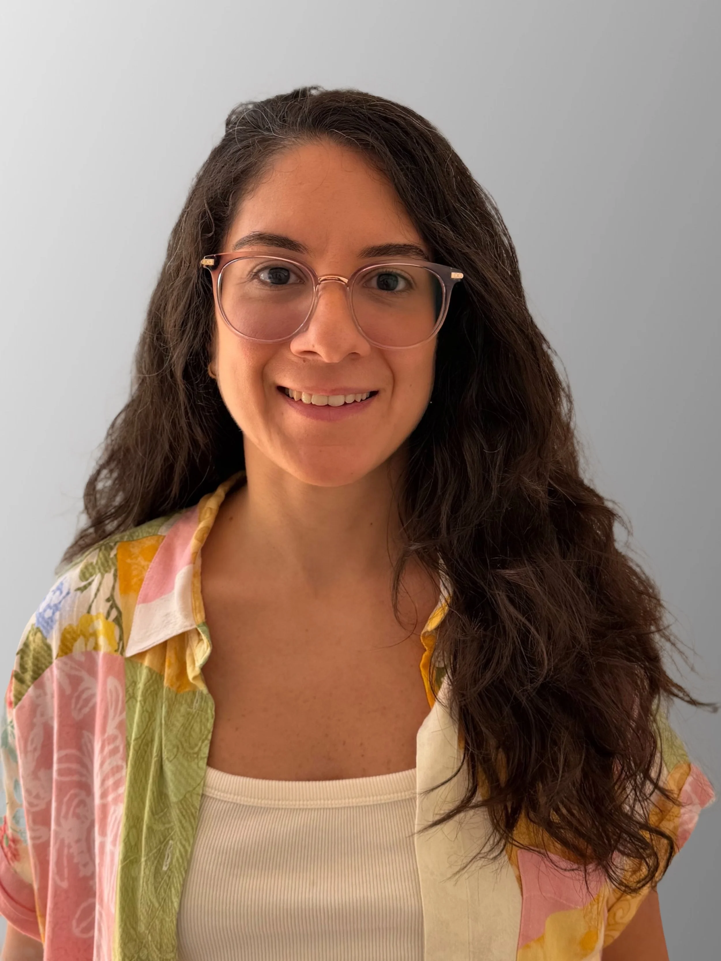 A woman with long, curly dark hair, wearing glasses with pink frames, a light-colored tank top, and a short-sleeved shirt with a colorful floral pattern. She is smiling and standing against a plain light gray background.