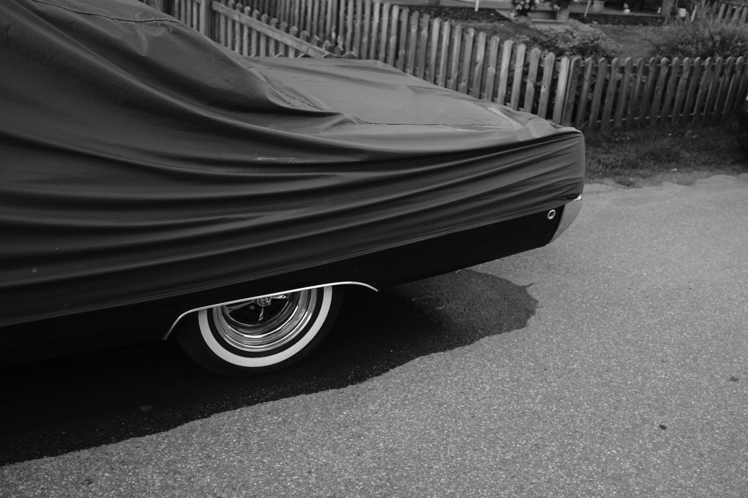 De achterkant van een auto die gedeeltelijk onder een zwarte afdekhoes is verborgen, met een witte wielrand zichtbaar en een kleine waterplas op de weg ernaast.