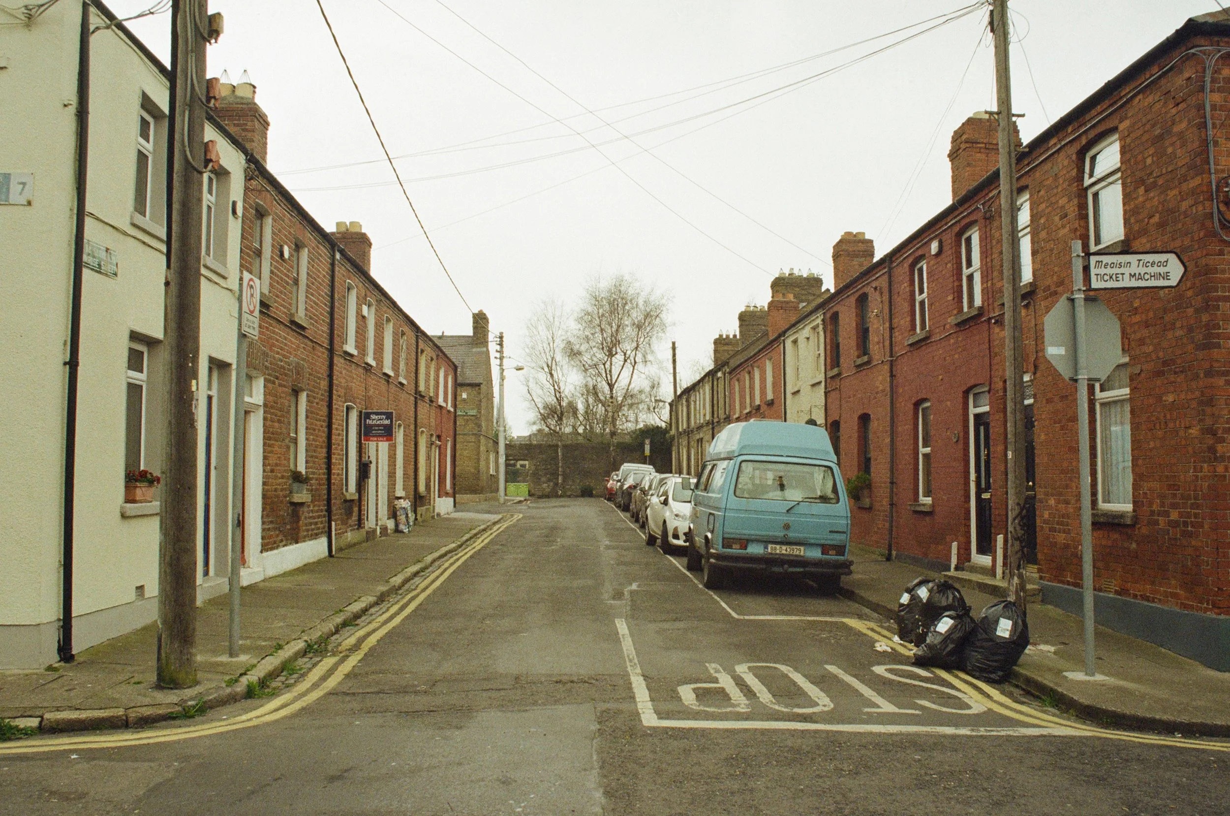 Een rustige straat met rijtjeshuis-woningen, parkeergelegenheid langs de weg en enkele vuilniszakken op de stoep, onder een grijze hemel.