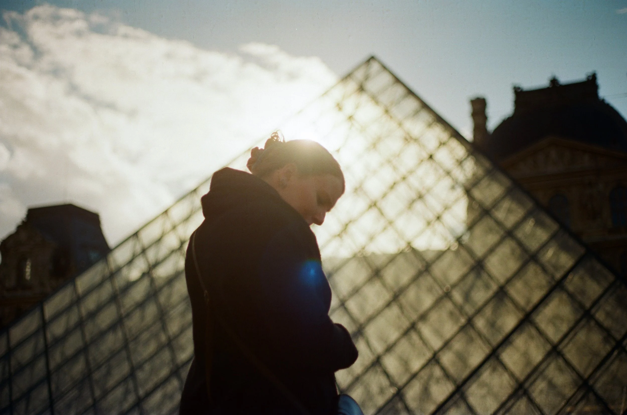 Een vrouw staat voor het Louvre in Parijs, haar silhouet zichtbaar tegen de zon, met het beroemde glasgewelfde museum op de achtergrond.
