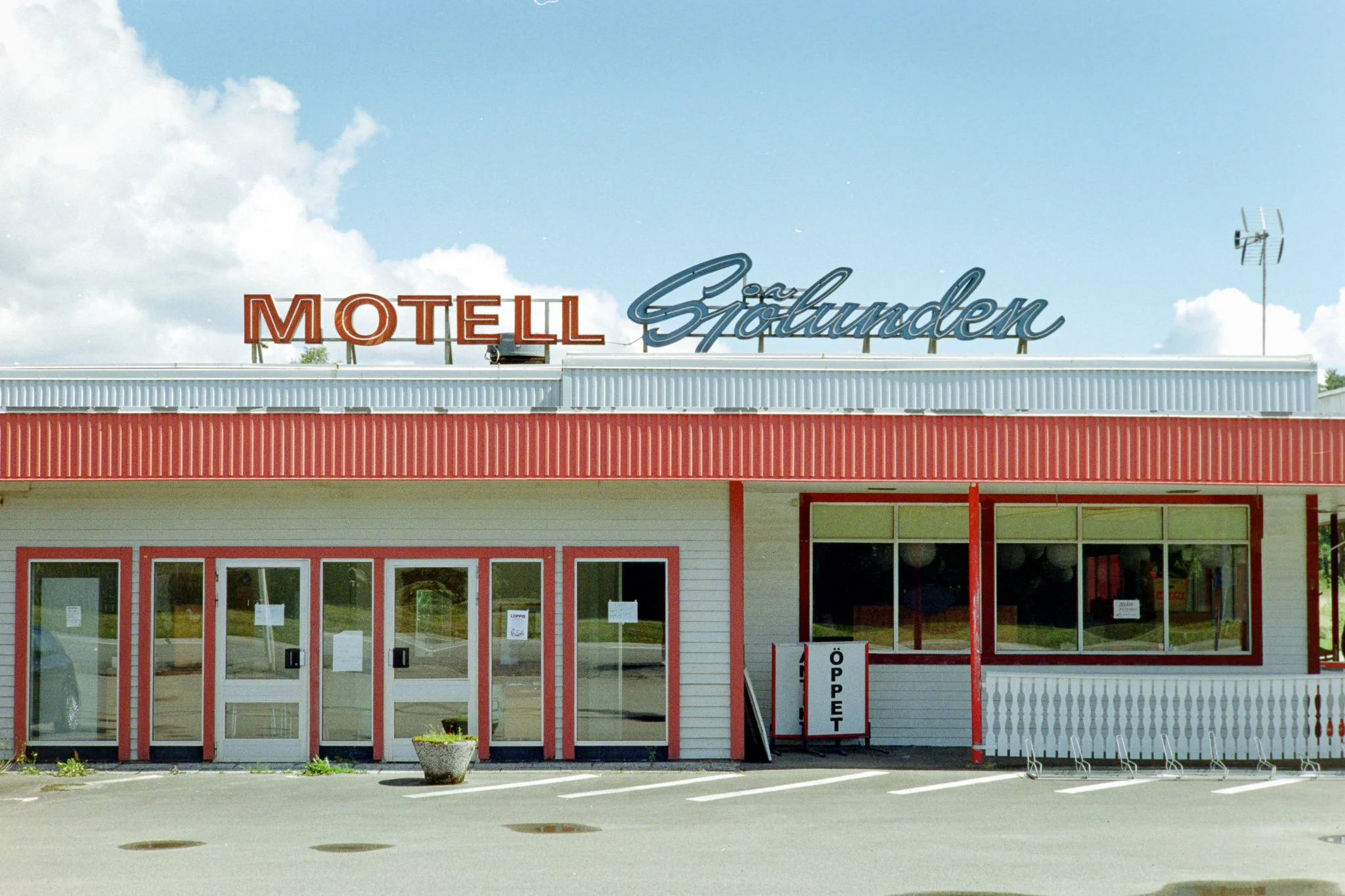 Een motel genaamd 'MOTELL' en 'Sjöland' met een wit en rood gebouw, parkeerplaatsen voor de ingang, een wit bord met 'ÖPPET' en grote ramen.