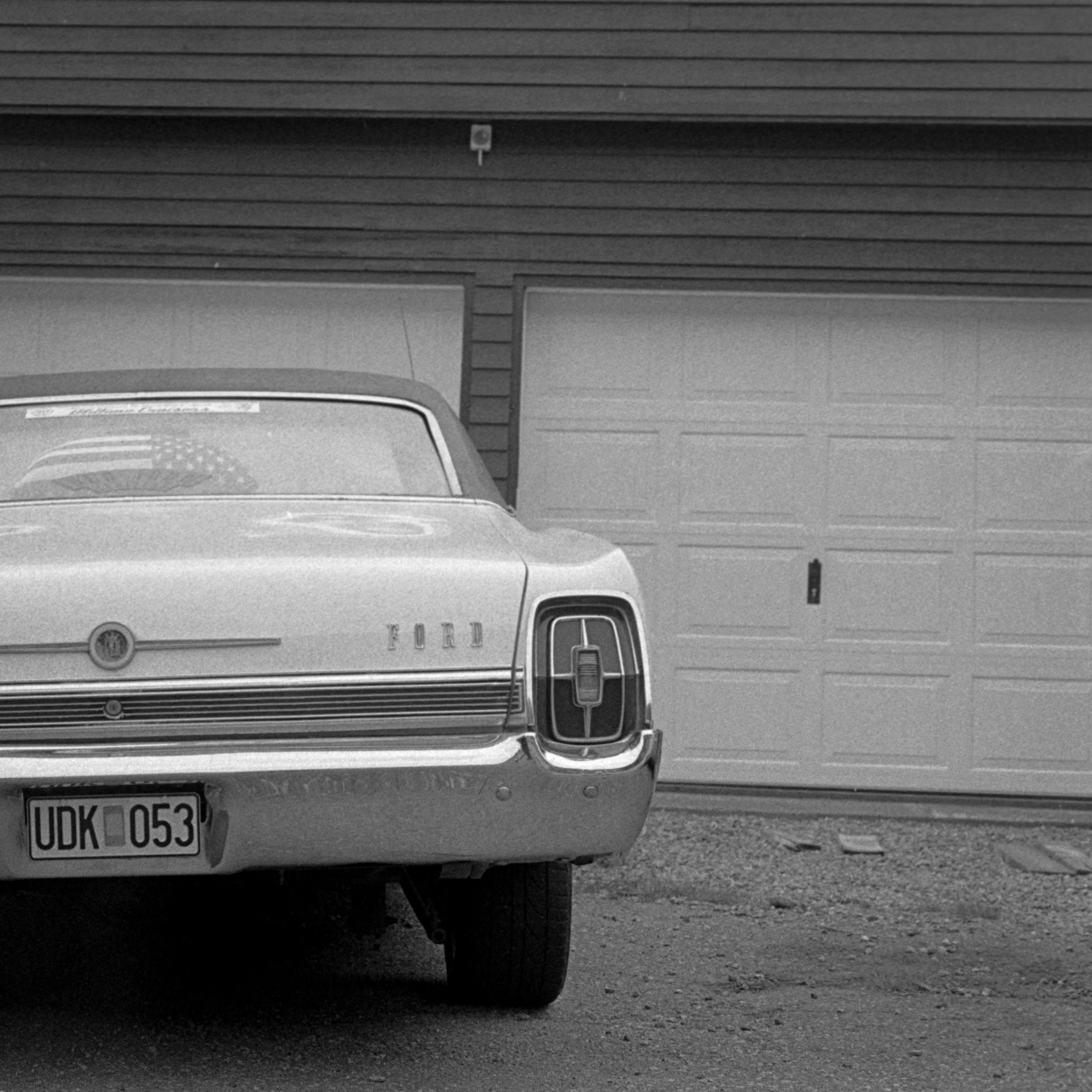 Een zwart-witte foto van een klassieke autootje voor een garage, met een Amerikaanse vlag op de achterkant. De kentekenplaat leest 'UDK 053'.