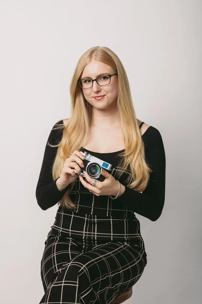 A woman with long blonde hair, wearing glasses, black top, and checkered pants, sitting on a stool, holding a vintage camera, and smiling at the camera.