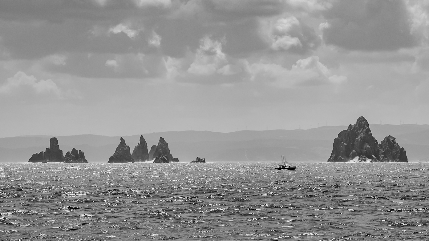 Vista del mar con islas rocosas y un barco pequeño navegando en el agua, con nubes en el cielo en una imagen en escala de grises.