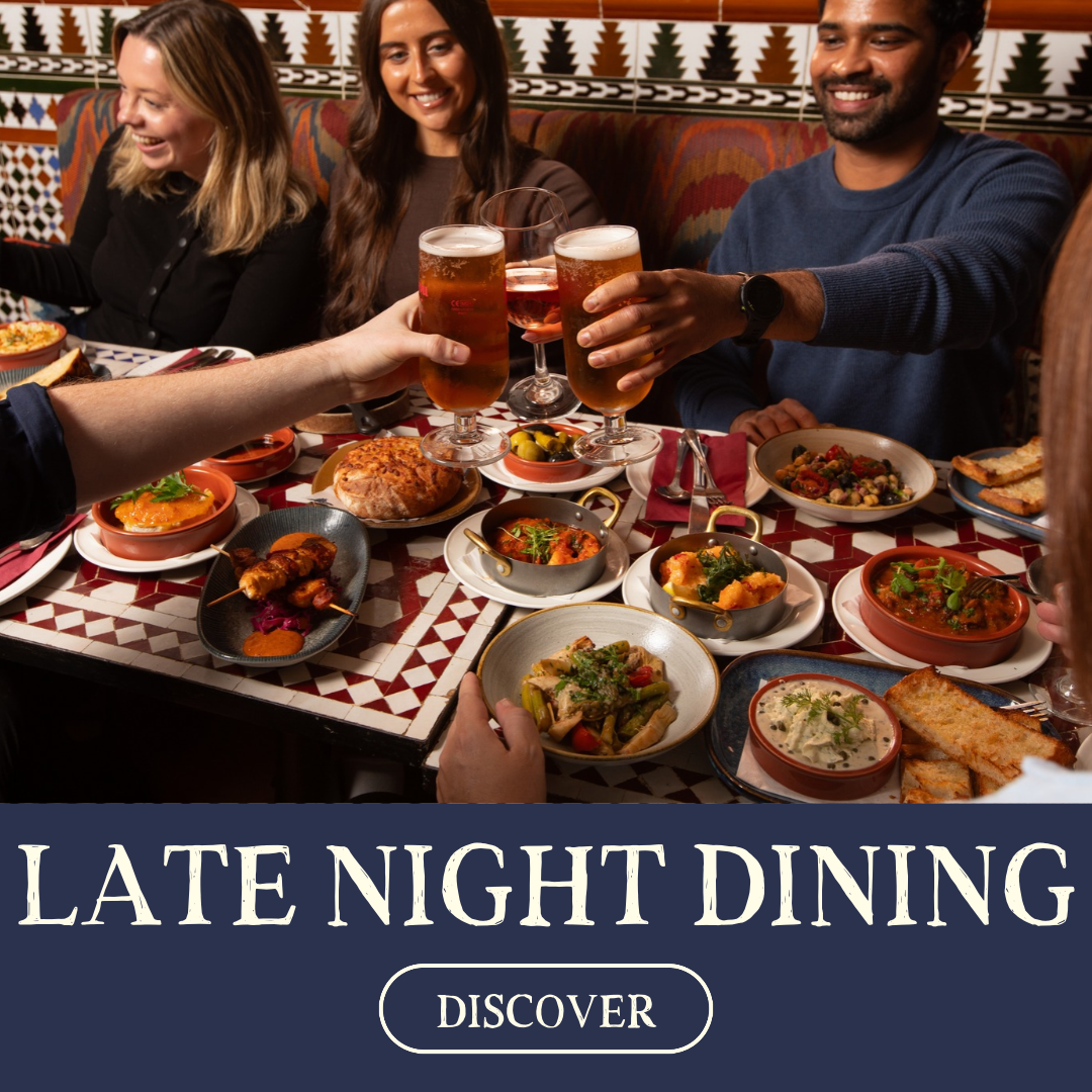 A group of friends enjoying late night dining at a restaurant, sharing drinks and a variety of dishes including soups, salads, and main courses.