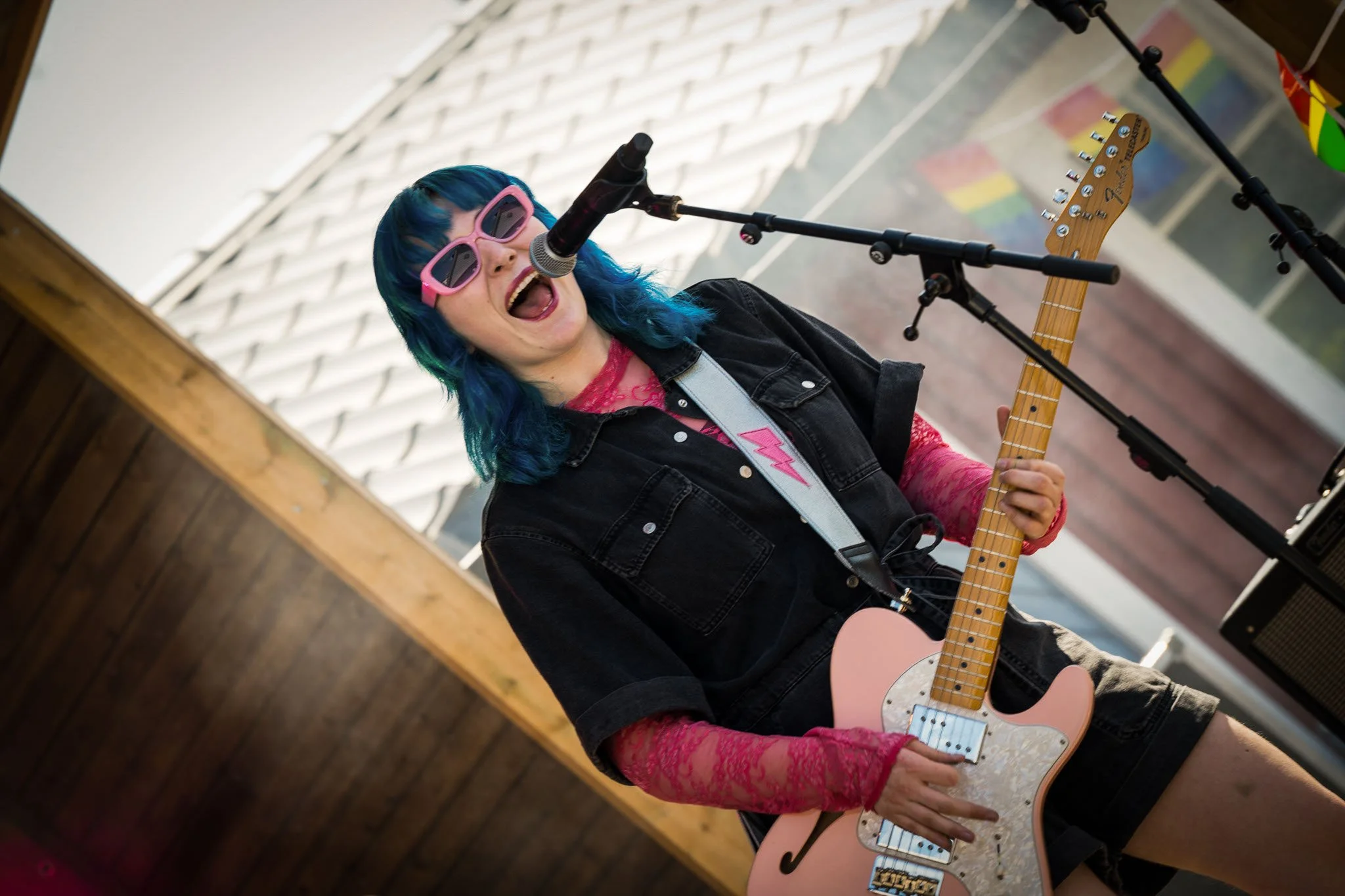 En kvinne med blått hår, rosa solbriller og svart denimjakke spiller elektrisk gitar mens hun synger inn i en mikrofon. Hun er på en scene med tre mikrofonstativ rundt seg.