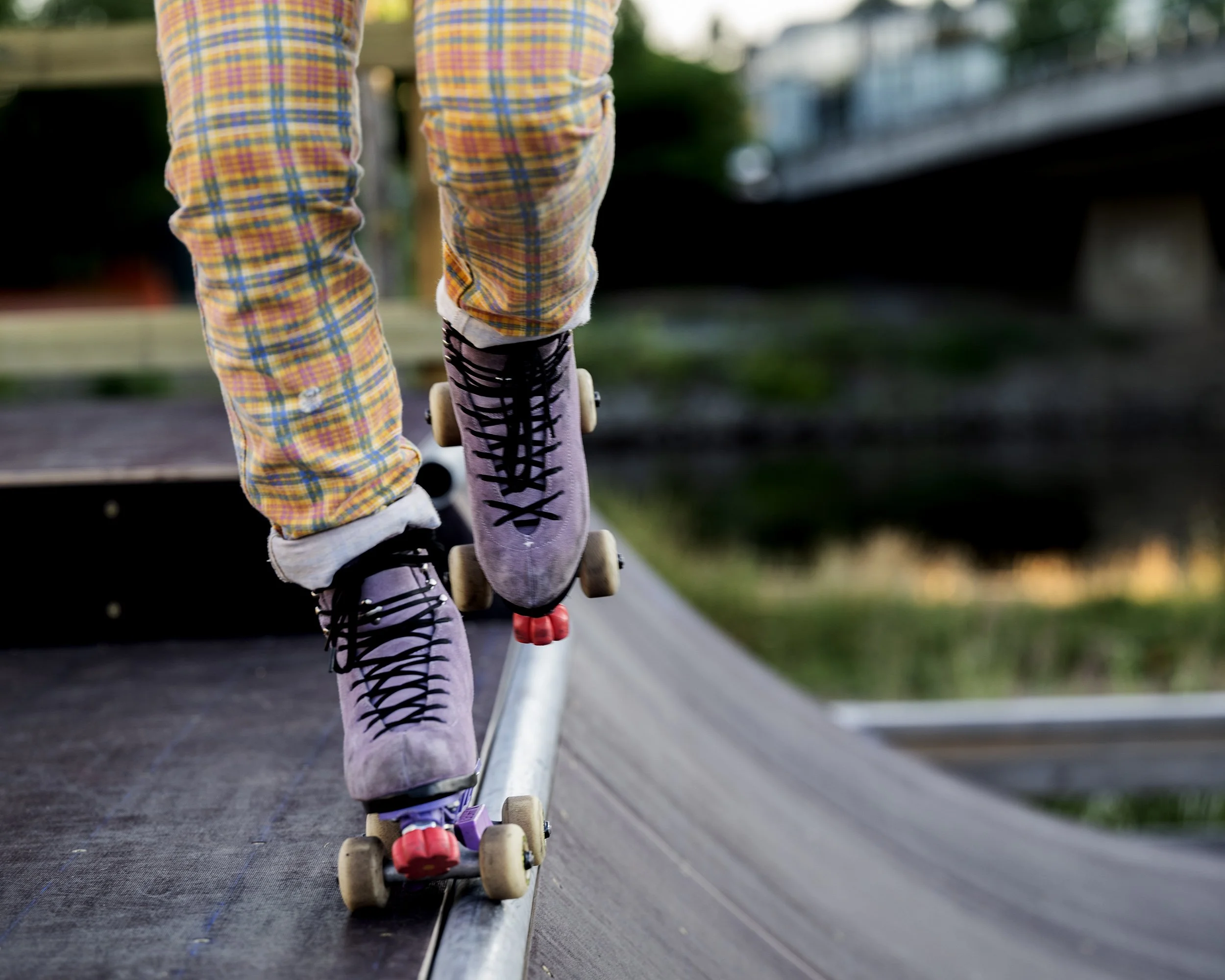 Person som bruker skateboard med lilla skatesko med svarte snører, og bukser med rutemønster, på en skateskatingbane.