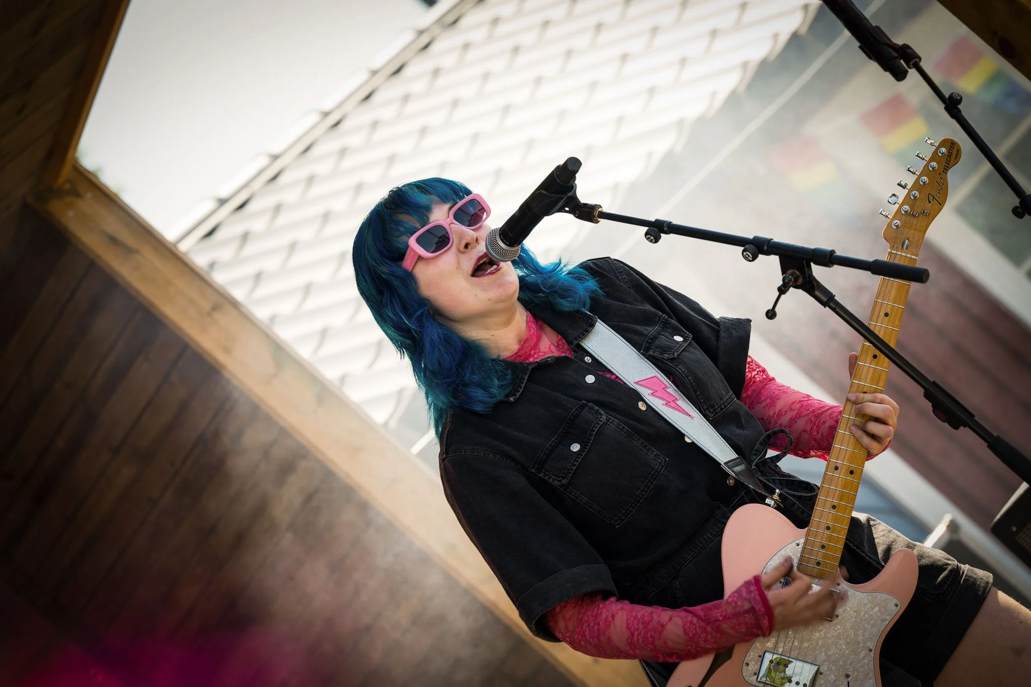 En kvinne med blå hår og rosa solbriller spiller gitar og synger inn i en mikrofon under en utendørs concert.