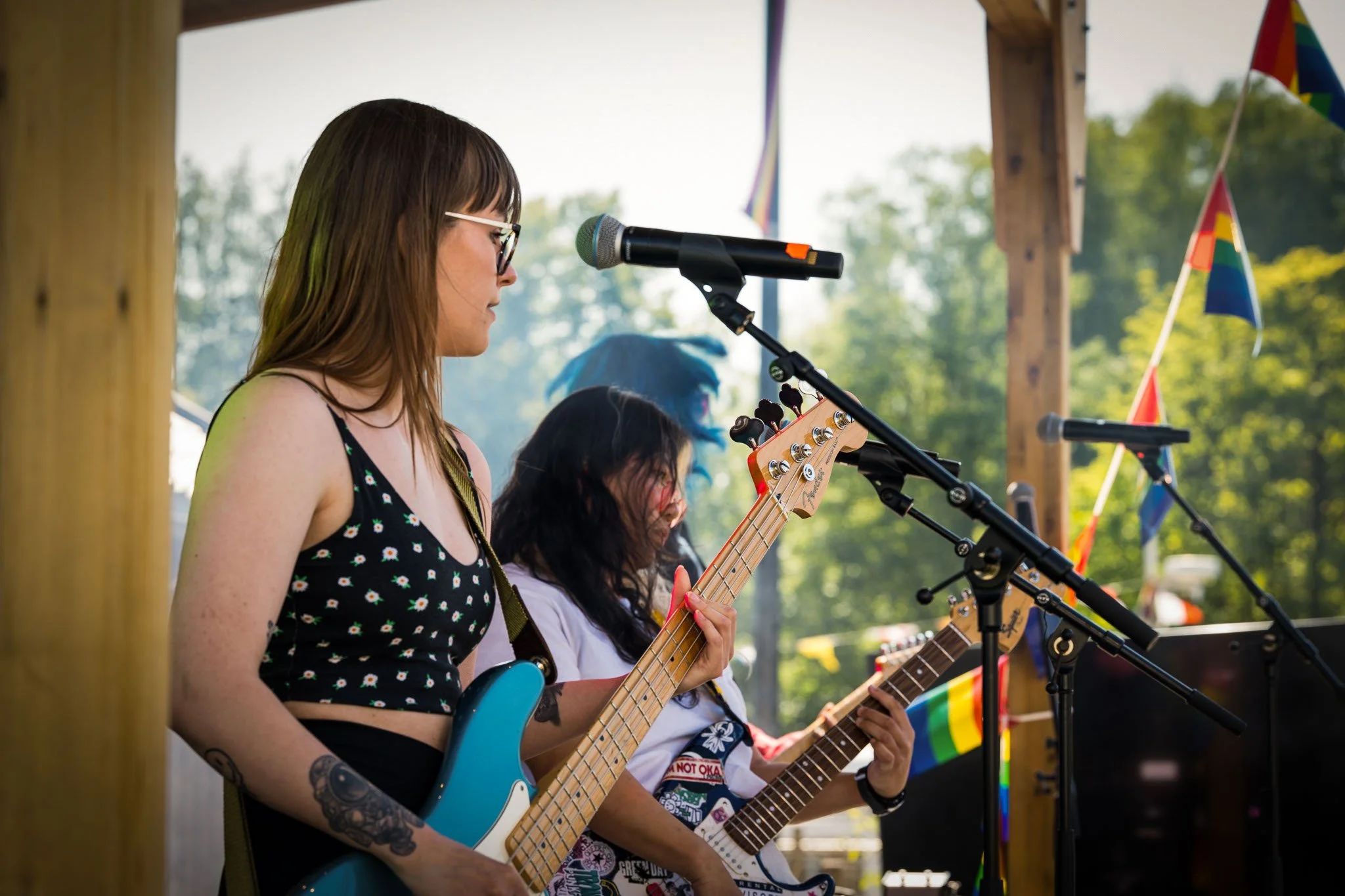 To kvinner spiller gitar på en utendørs scene dekket av tre med små regnbue- og pride-flagg. Den ene kvinnens sideprofil er synlig, hun har brunt hår, briller og tatoveringer på armen.
