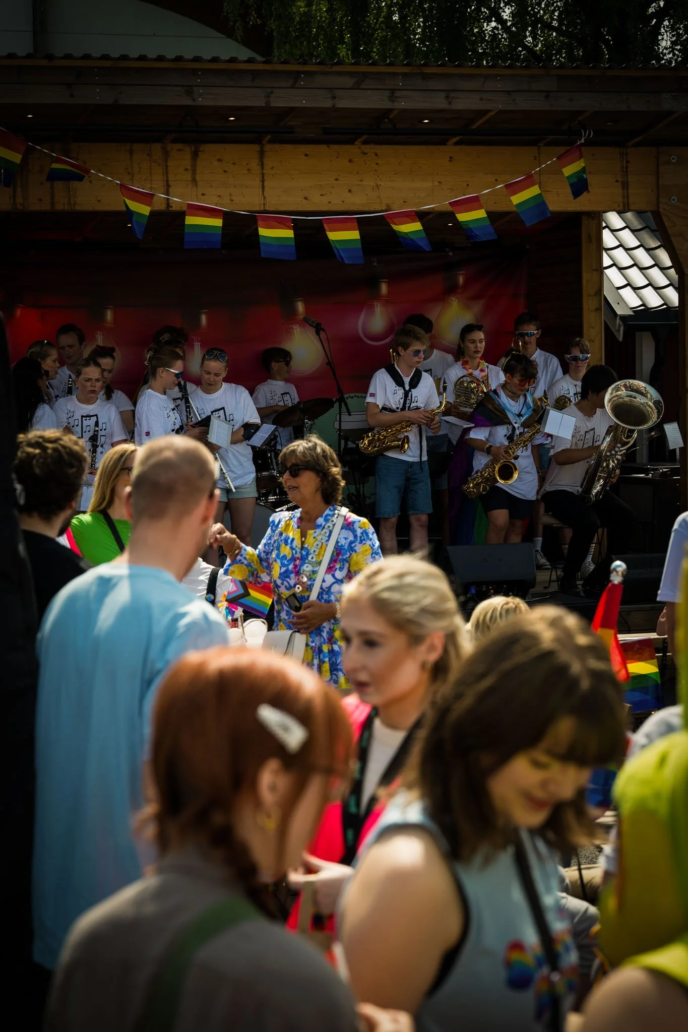 En gruppe mennesker som feirer utendørs under en Pride-parade, med et band som spiller på scenen i bakgrunnen og Pride-flagg og bannere som pynter området.