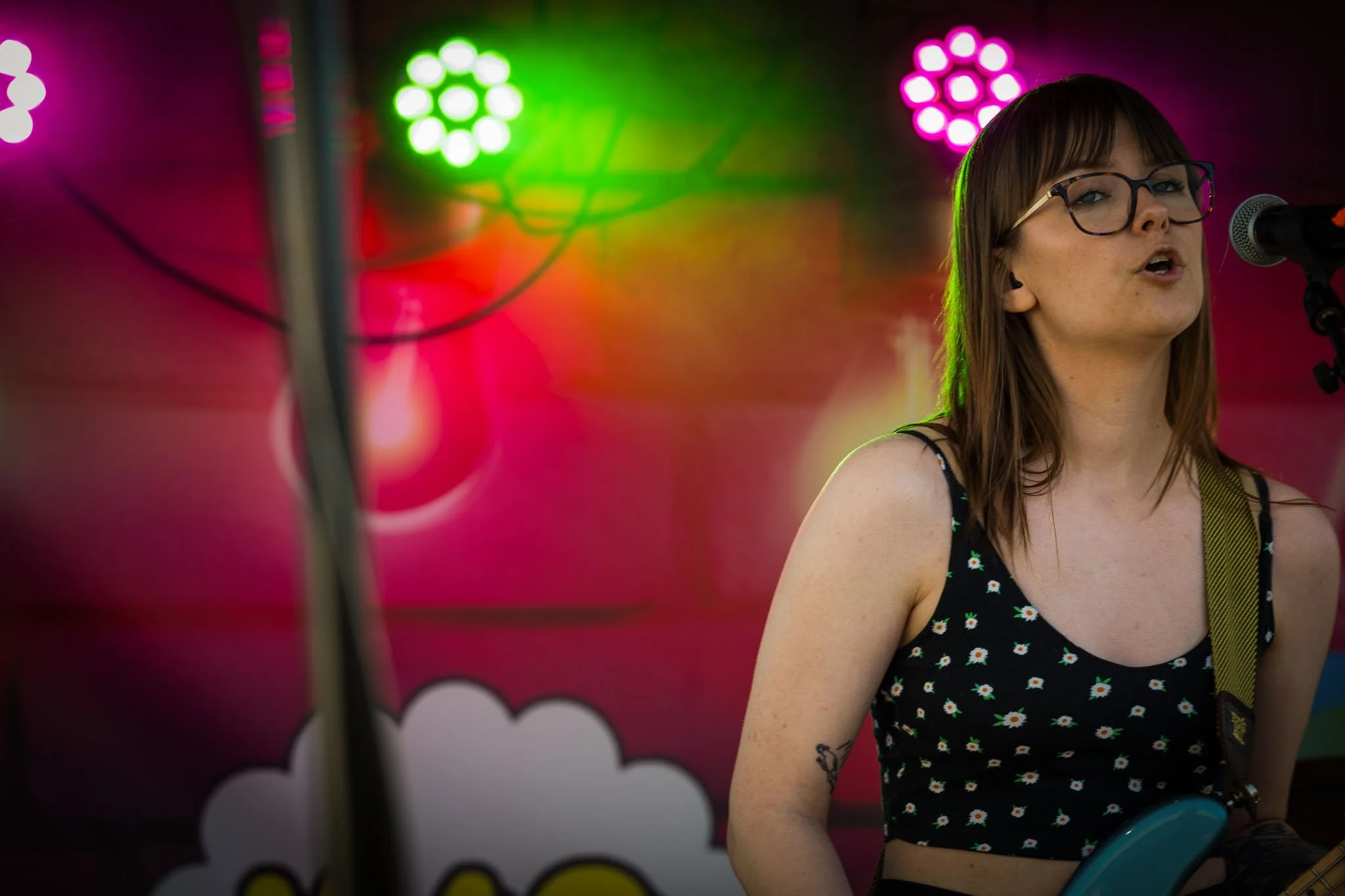 En kvinne med briller spiller bassgitar på scenen med fargerike neonlys i bakgrunnen.