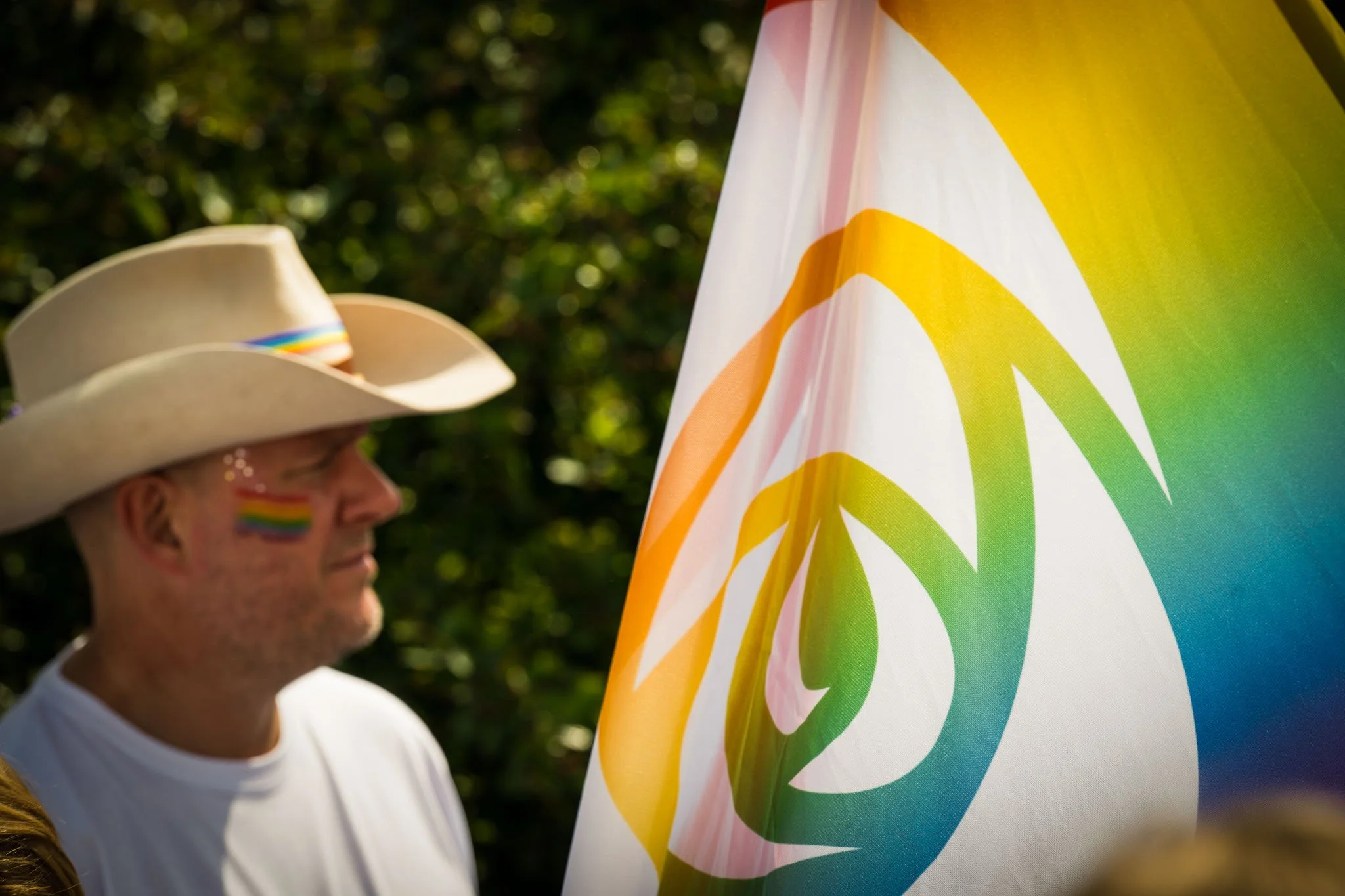 Person med regnbuefargede flaggmerker i ansiktet, iført stor hat, står nær en fargerik regnbuefarget flagg.