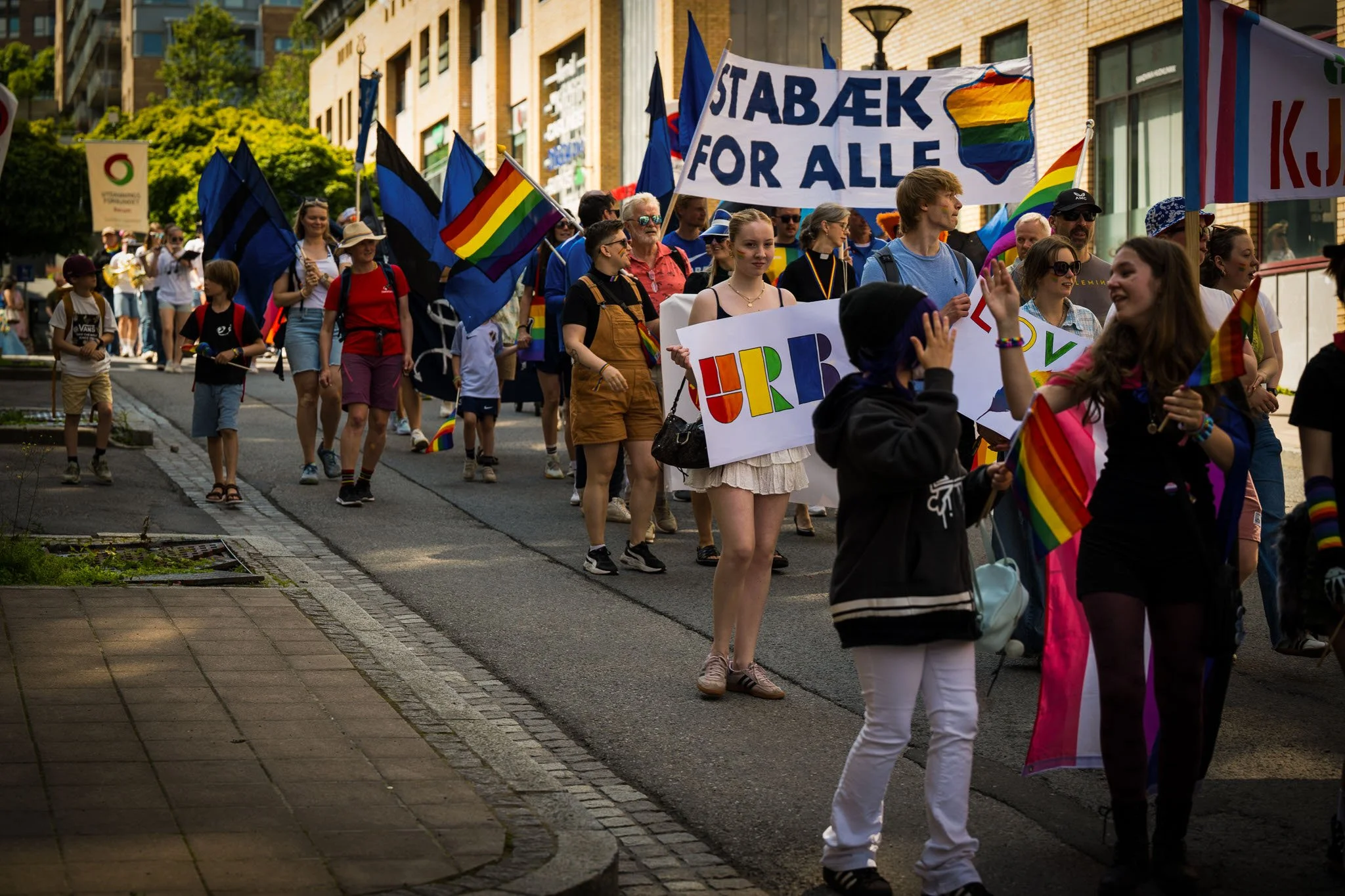 Folk deltar i en parademarsj med regnbueflag, tegn med tekster som "Stabæk for alle" og "URB" i en bygate.
