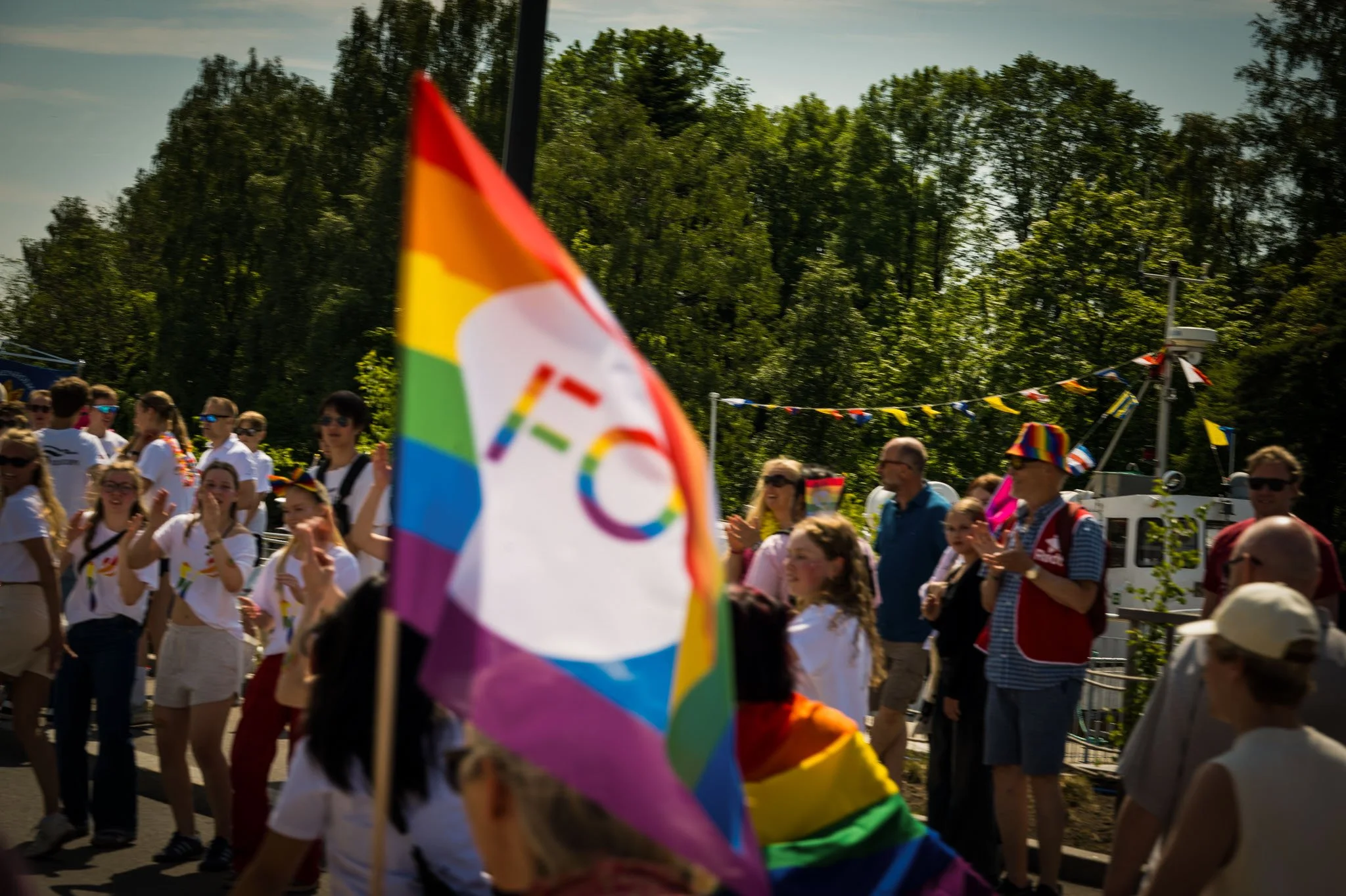 Folk som deltar i en Pride-parade med rainbow-flagg i solskinn, mange smiler og heiser hender, med trær og en karusell i bakgrunnen.