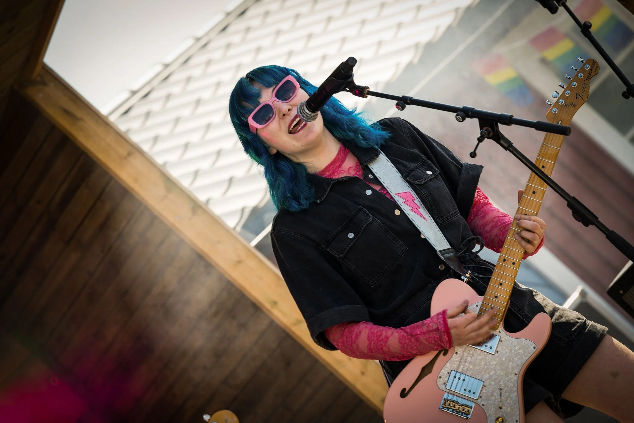 Kvinne med blått hår, rosa solbriller, i svart jakke og rosa netting overdel, spiller gitar og synger inn i mikrofon.
