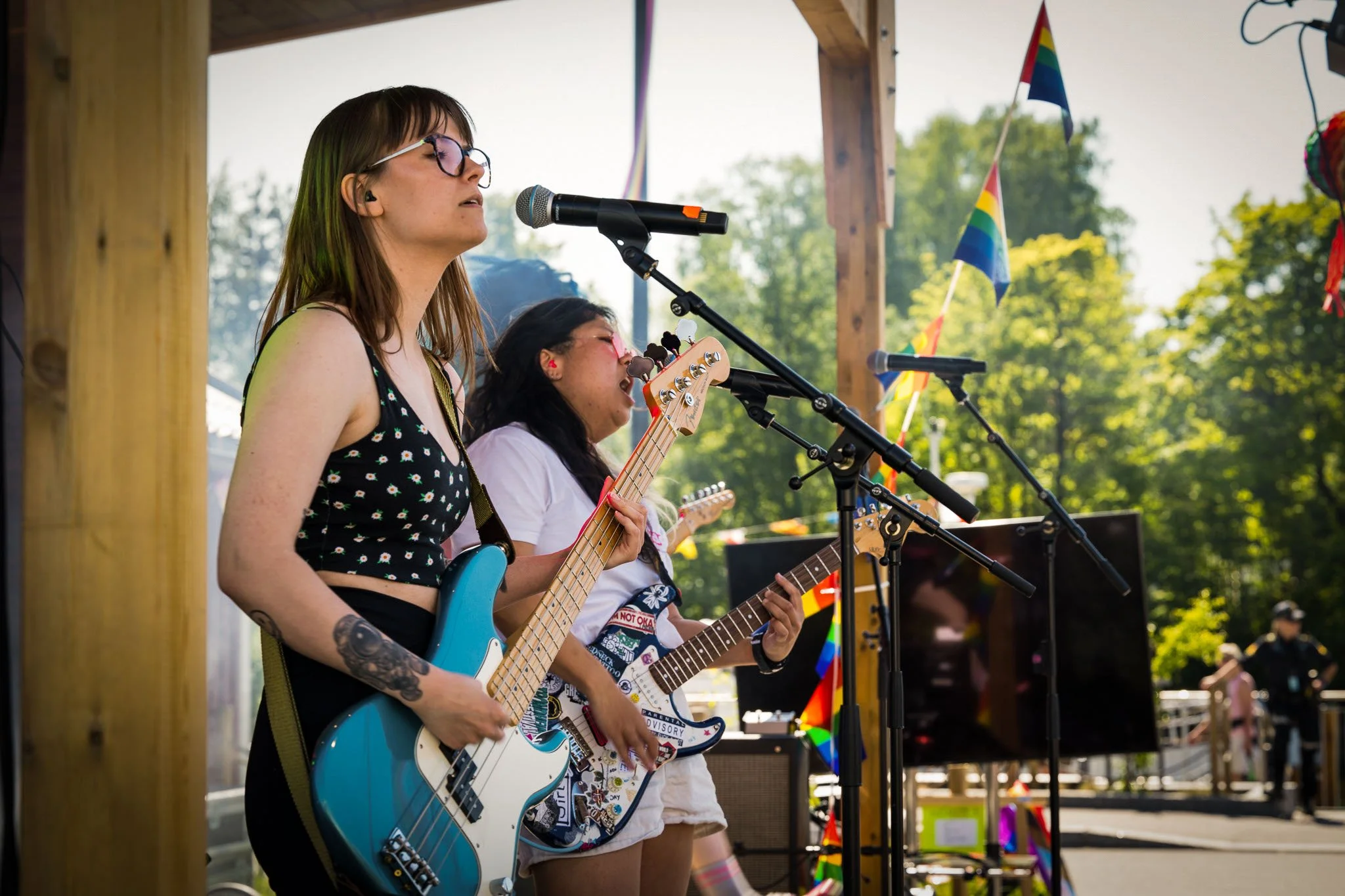 To kvinner spiller gitar og synger på en scene utendørs, med regnbueflagg og grønne trær i bakgrunnen.