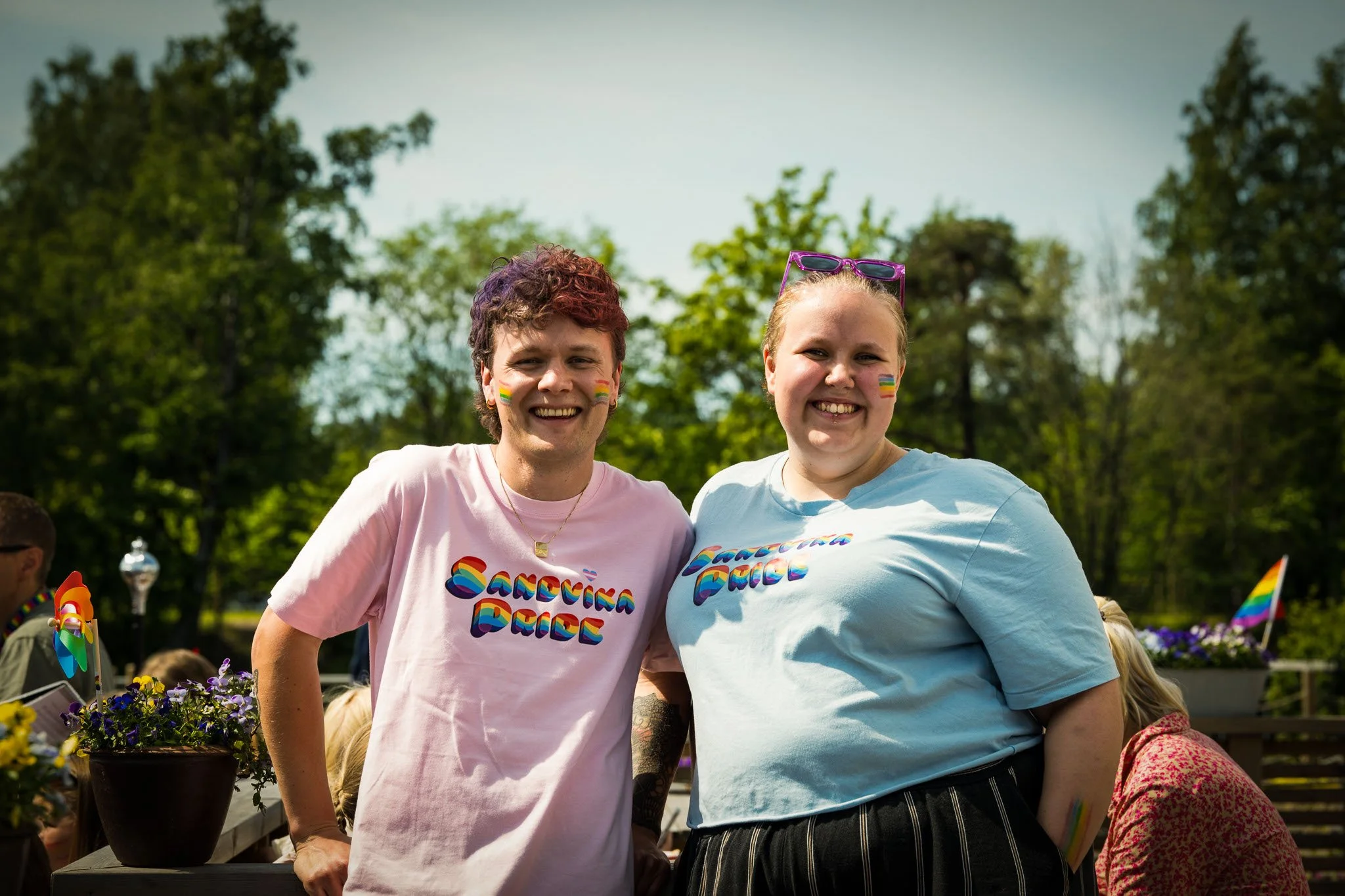 To personer smiler under en utendørs pride-feiring, begge med påfargede runde flaggstikk på kinnene og T-skjorter med regnbuefarger og teksten 'Candy Girl' og 'Candy Boy'.