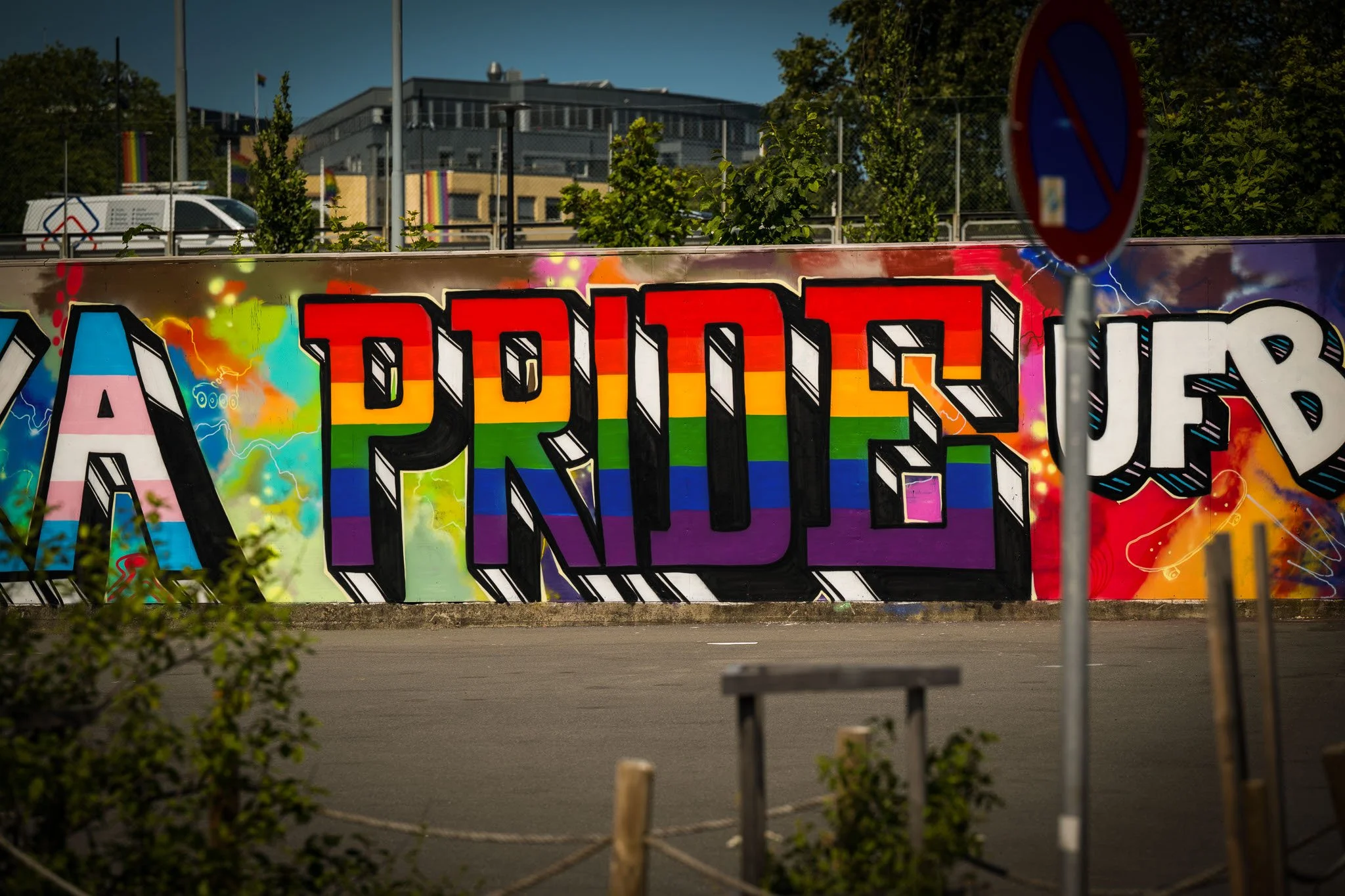 Graffiti med teksten "A PRIDE" i regnbuefarger på en vegg, omgitt av grønne busker og trær, med en stang med forbudsskilt foran.
